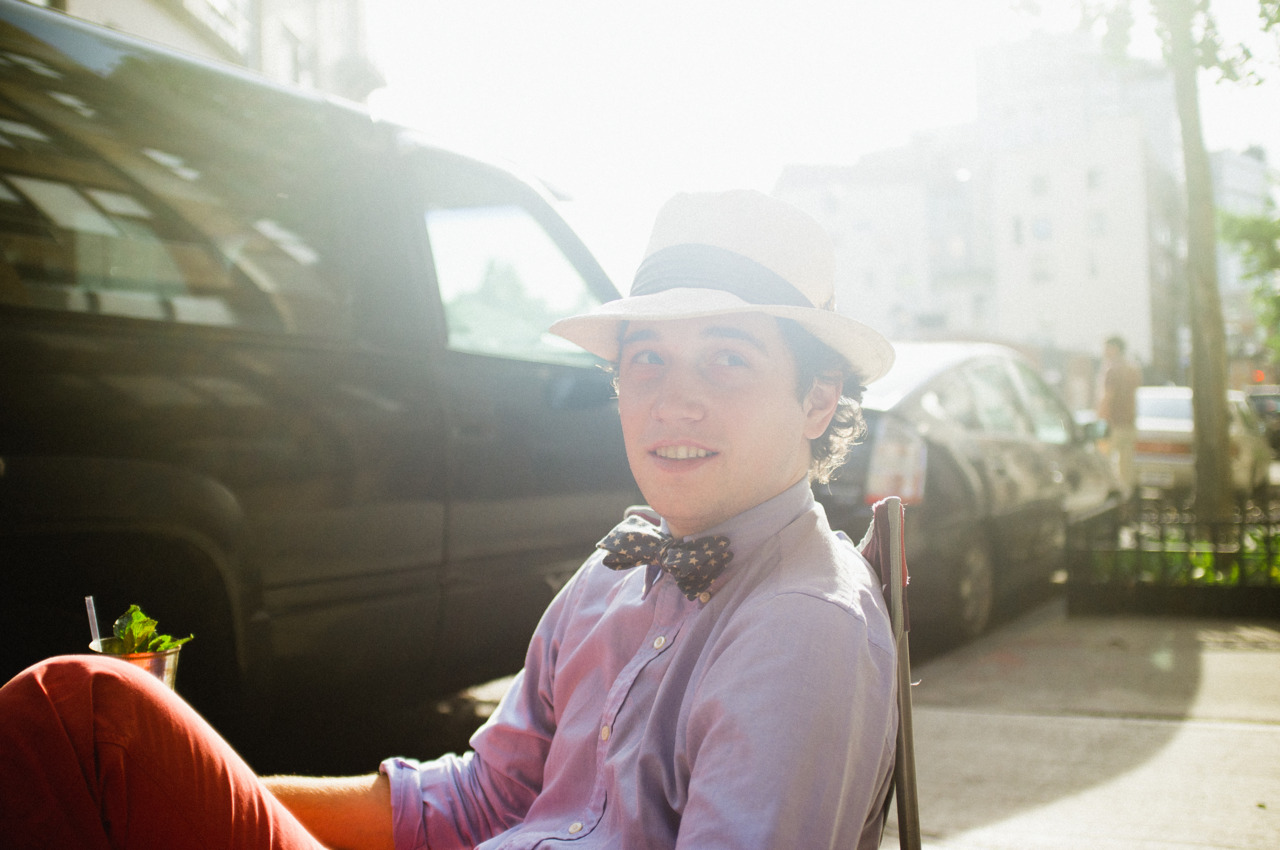 Smiling young man in a straw fedora and patterned bow tie seated outdoors on a sidewalk folding chair.