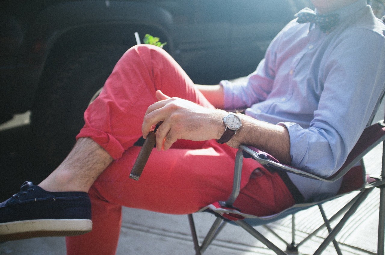 Man in red trousers and a gingham shirt holding a cigar while relaxing outdoors in the sunlight.