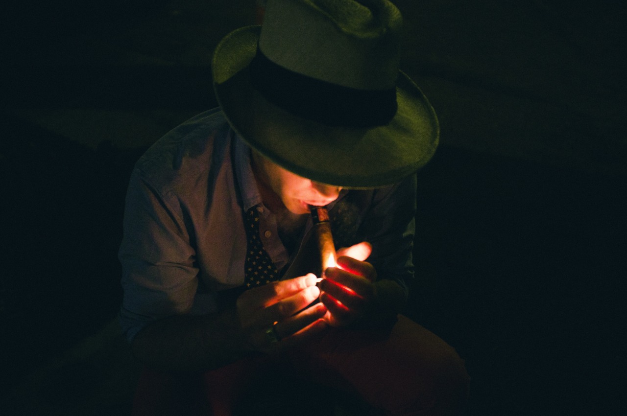 Man wearing a fedora lighting a cigar in the dark with bright flame glow illuminating his face.