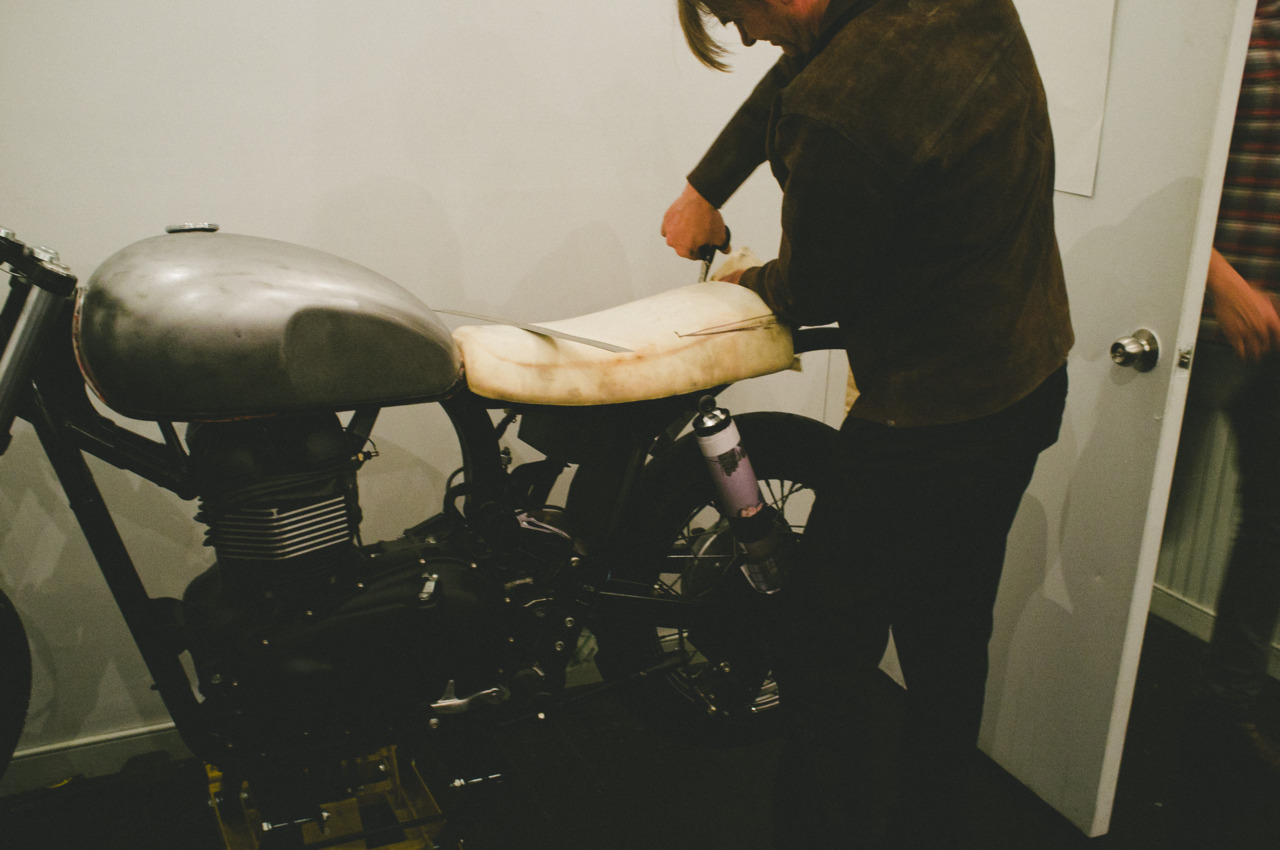 Craftsman shaping a custom foam motorcycle seat onto a stripped cafe racer frame in a workshop setting.