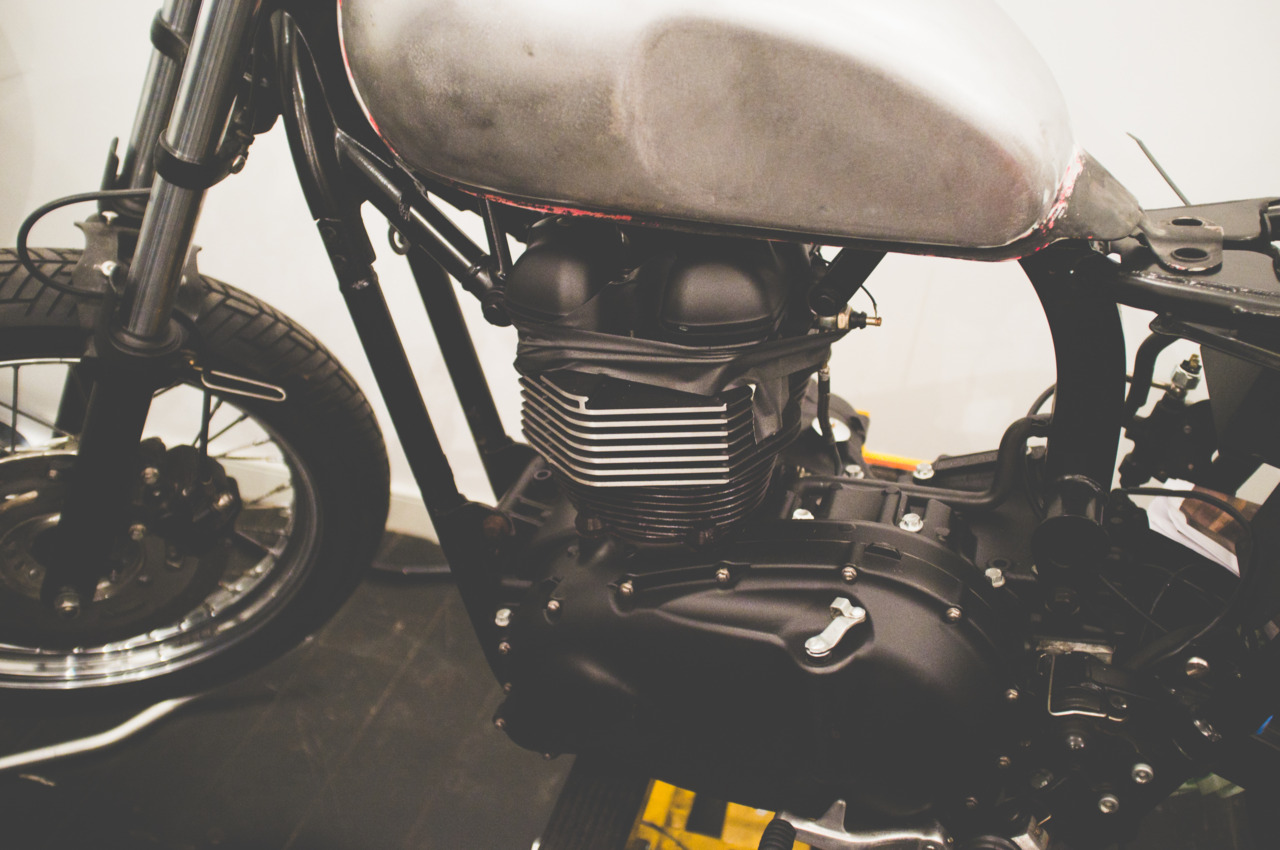 Raw brushed steel fuel tank mounted above an exposed twin-cylinder motorcycle engine on a custom build.