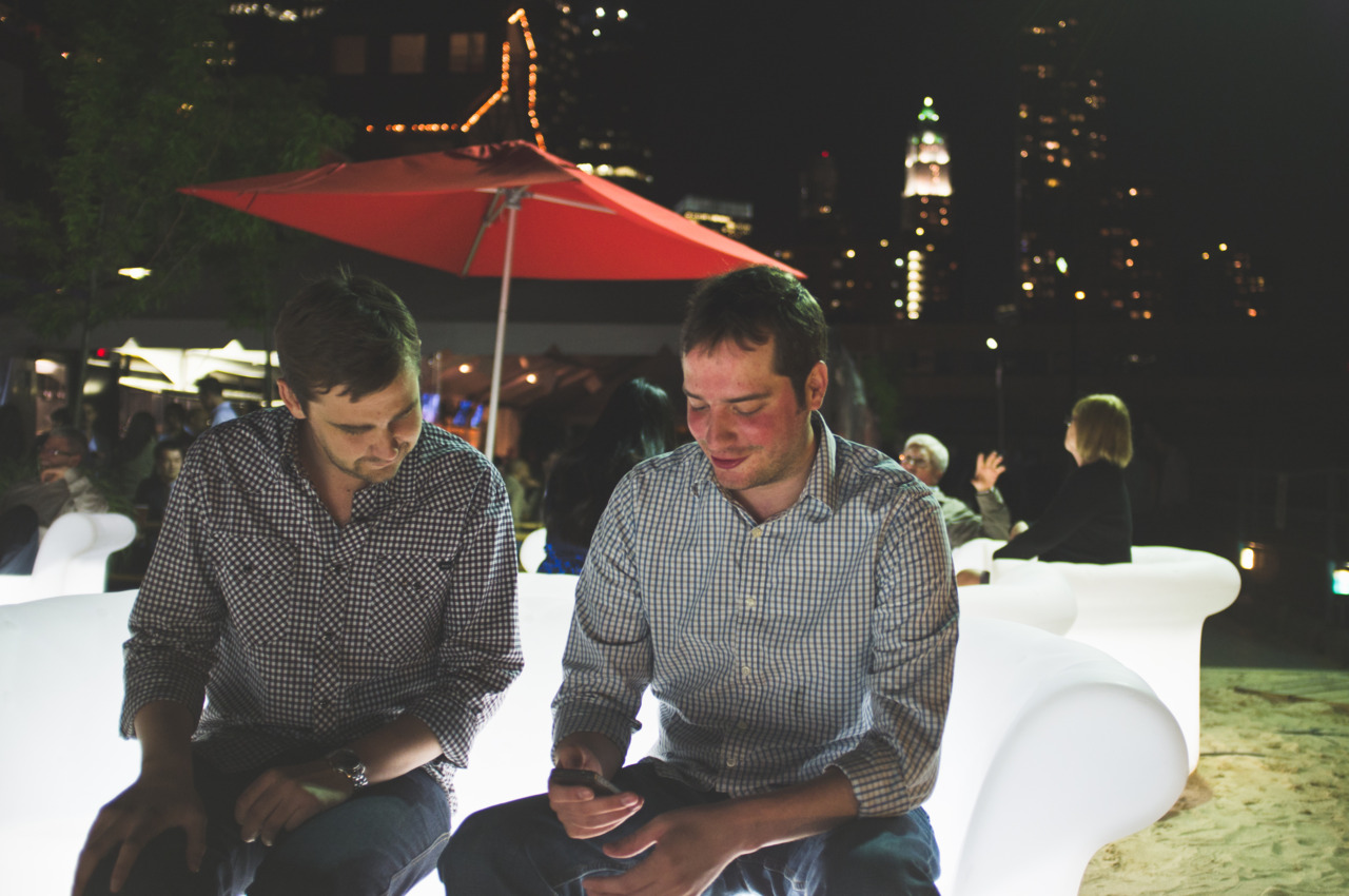 Two men in collared shirts seated on an illuminated curved bench checking a phone at Beekman Beer Garden at night.