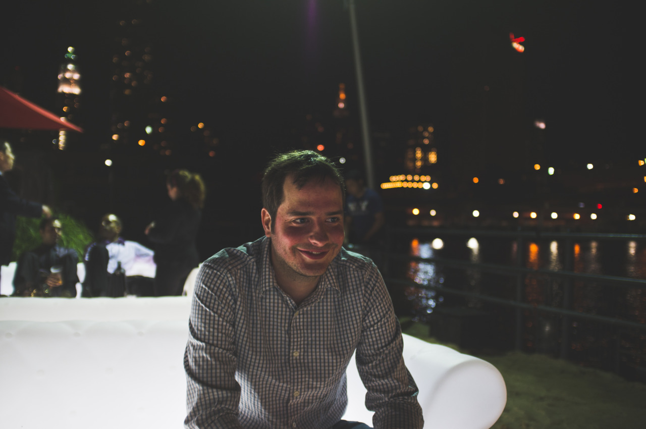 Smiling man in a gingham shirt on an illuminated bench with waterfront lights at Beekman Beer Garden.