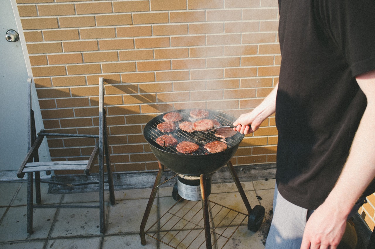 Man grilling hamburger patties with a spatula on a round charcoal grill during an outdoor rooftop barbecue.