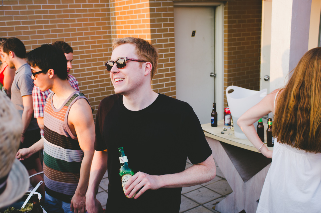 Smiling man wearing sunglasses holding a beer bottle while chatting with friends at a rooftop party.