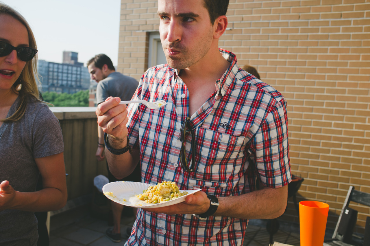 Man in a plaid short-sleeve shirt eating rice off a paper plate at an outdoor patio cookout.