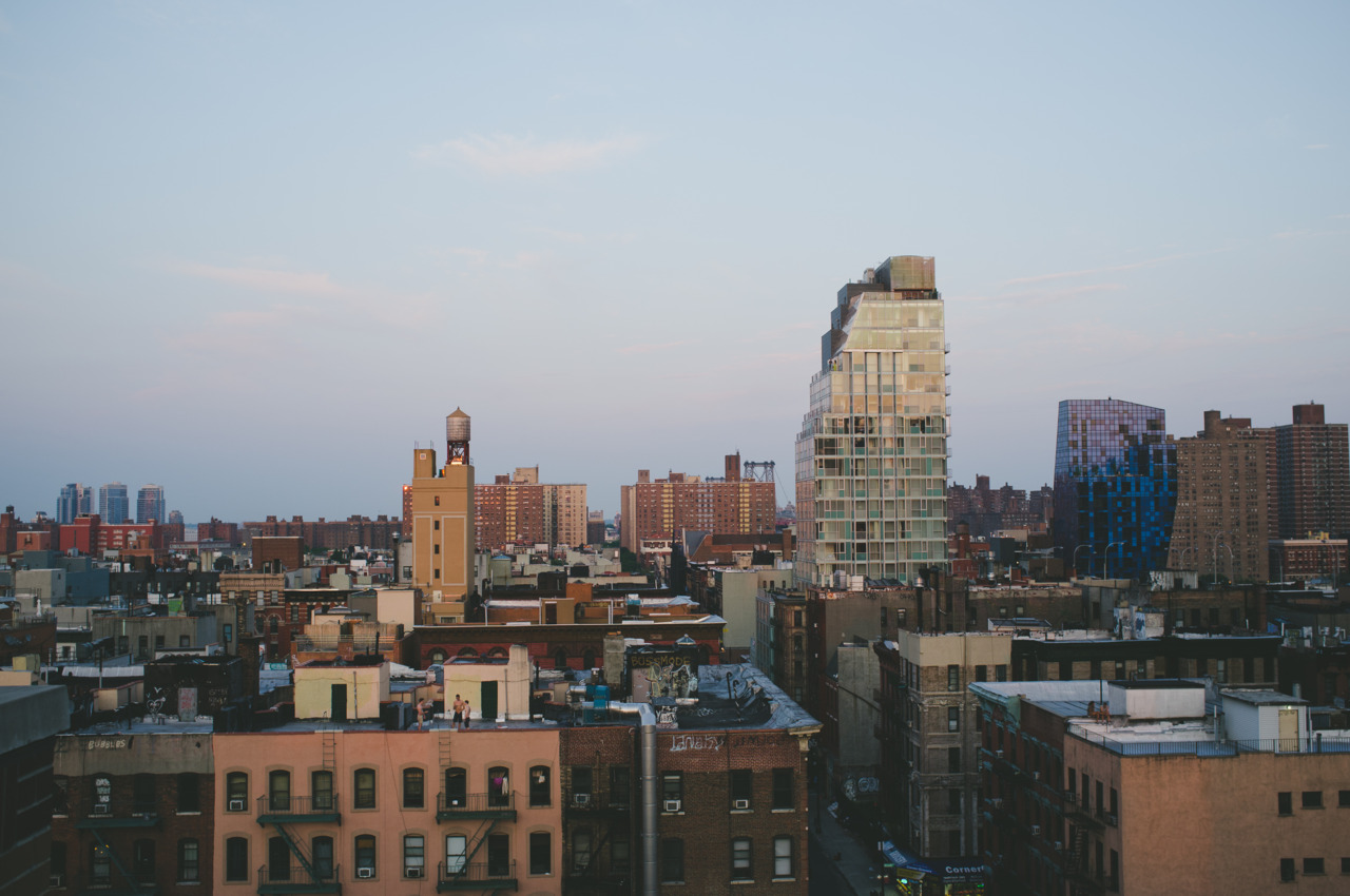 Rooftop view of brick tenement buildings and water towers against modern glass high-rises at sunset.