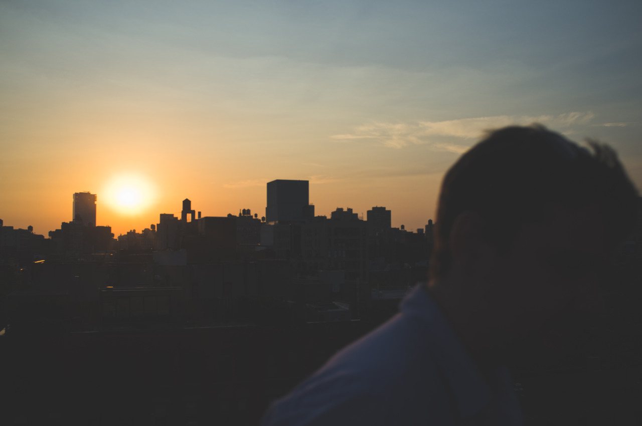 Silhouetted profile of a person looking out over urban rooftop water towers toward a golden sunset.