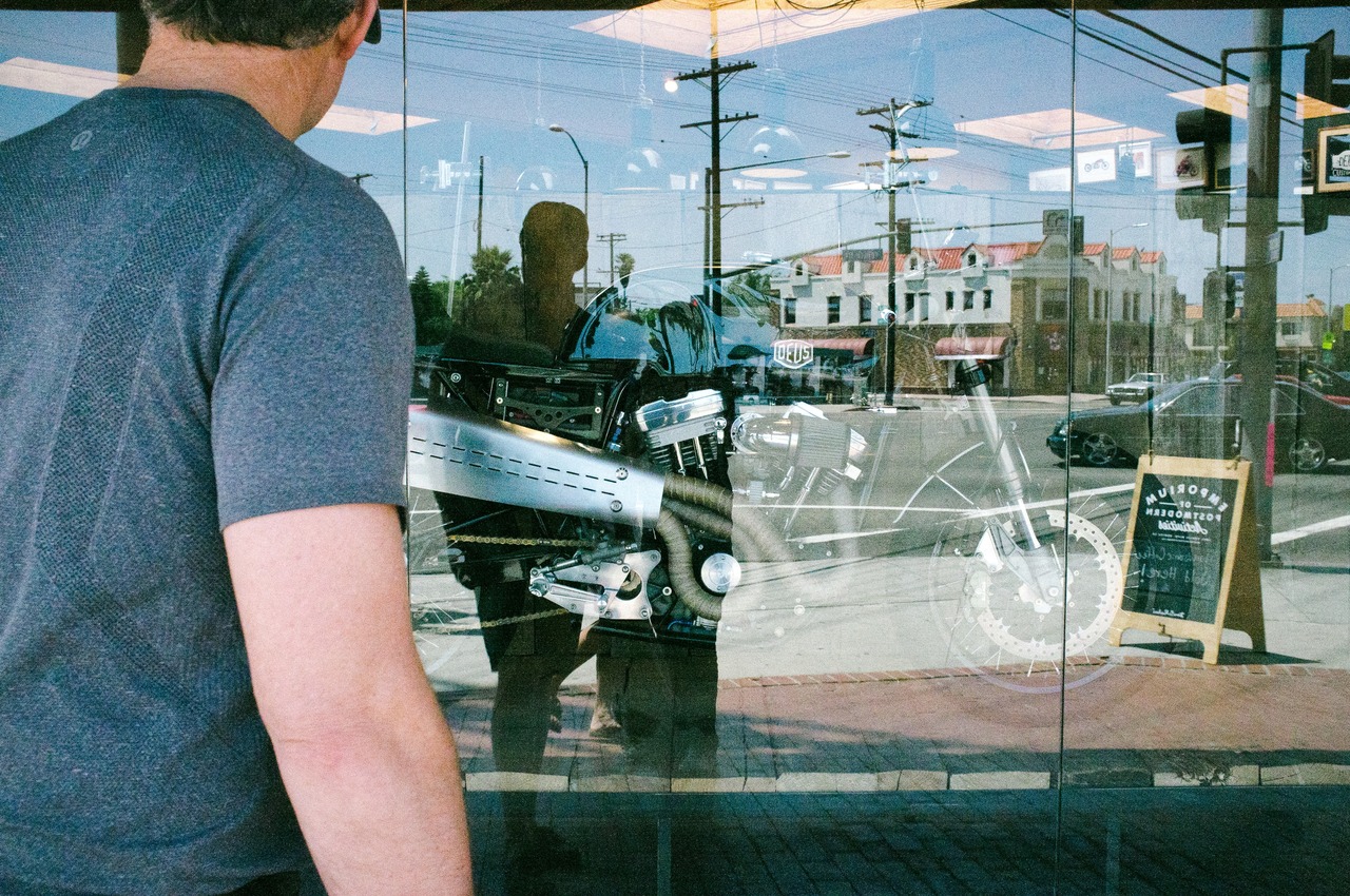 Over-the-shoulder view of a man looking through the glass storefront window of Deus Ex Machina at a custom motorcycle reflecting Venice street architecture, captured with a Fujifilm FinePix X100.