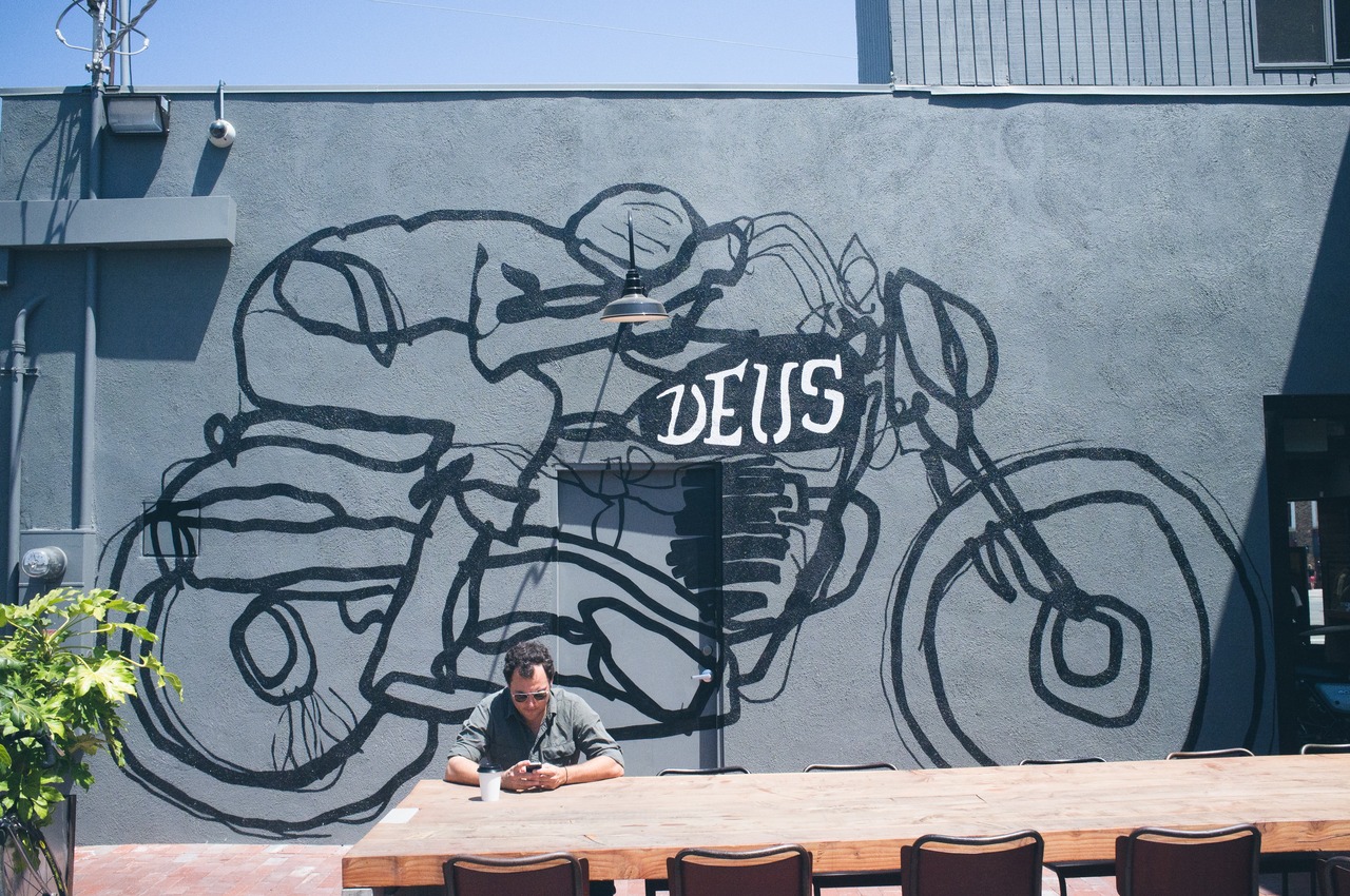 Man sitting at a long wooden communal outdoor table checking his phone beneath a large motorcycle racer wall mural at Deus Ex Machina in Venice, captured with a Fujifilm FinePix X100.