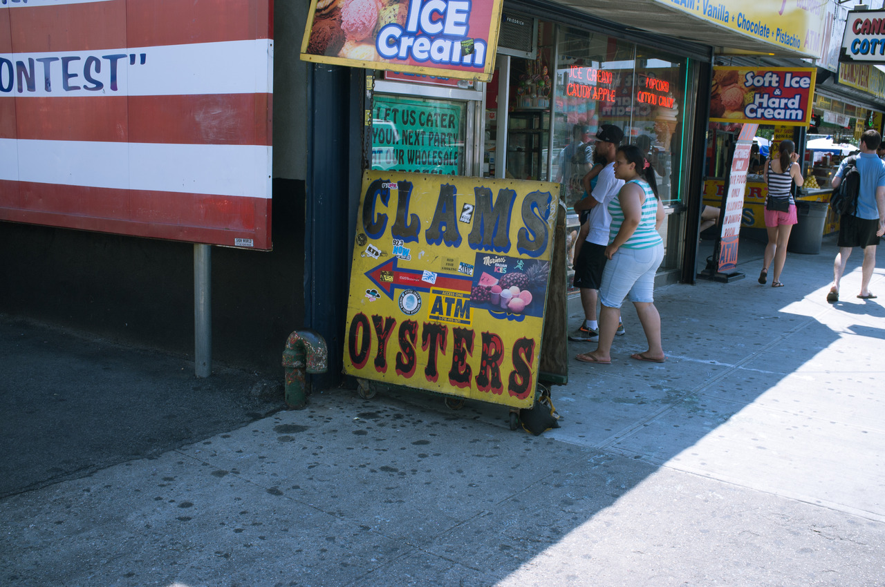 Yellow sandwich board sign advertising 'CLAMS' and 'OYSTERS' on the Coney Island boardwalk.