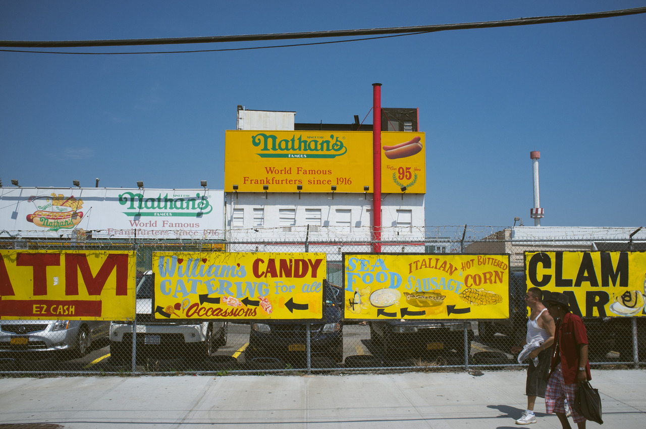 Pedestrians walking past yellow hand-painted food signs on a fence outside Nathan's in Coney Island.