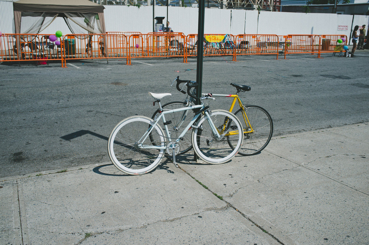 Two vintage road bicycles locked to a lamppost on a street with orange barricades near Coney Island.