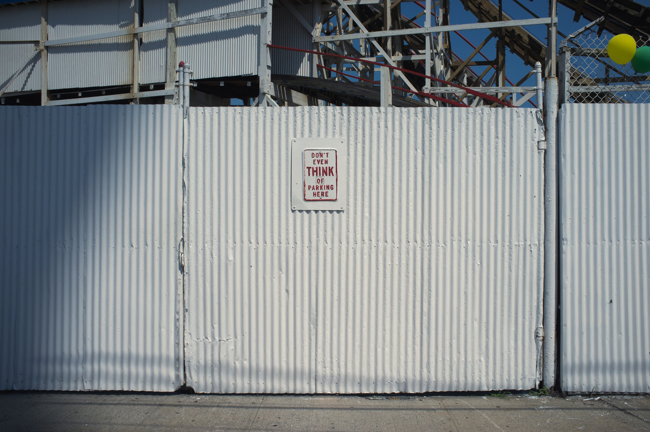 Corrugated metal wall beneath wooden rollercoaster trestles with a sign reading 'DON'T EVEN THINK OF PARKING HERE'.