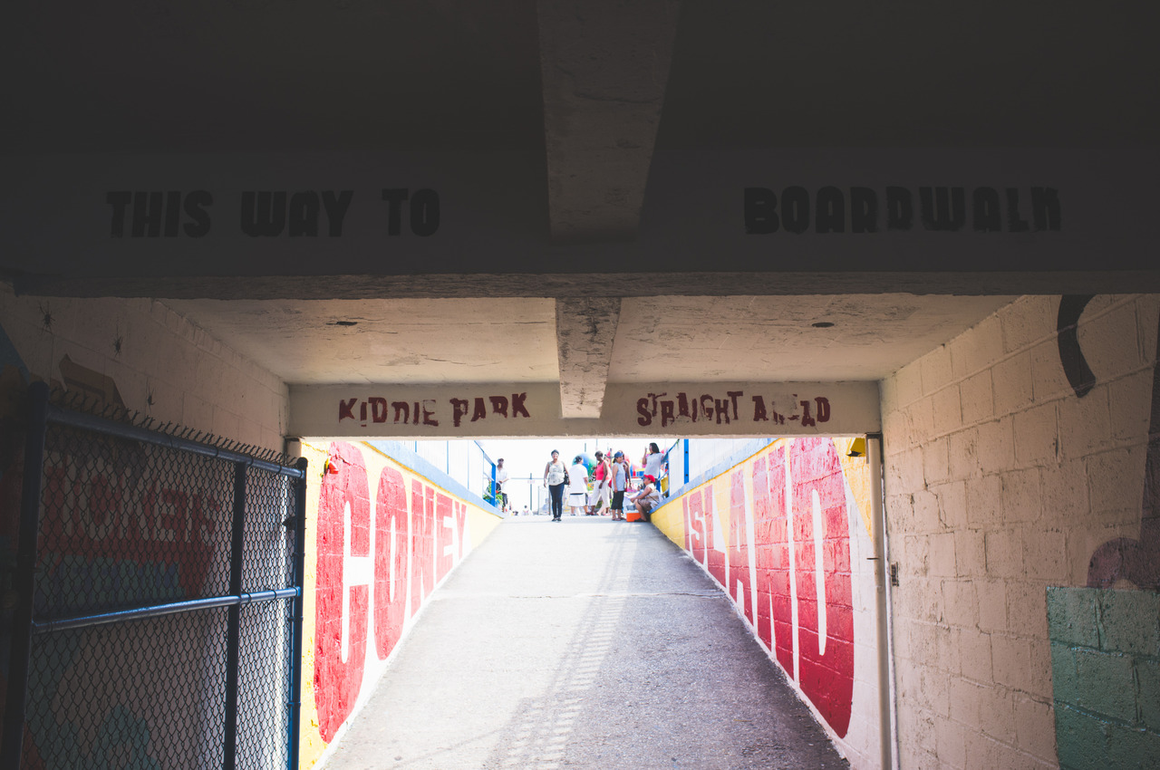 Pedestrian ramp tunnel toward the sunny boardwalk, lined with red 'CONEY ISLAND' brick lettering.