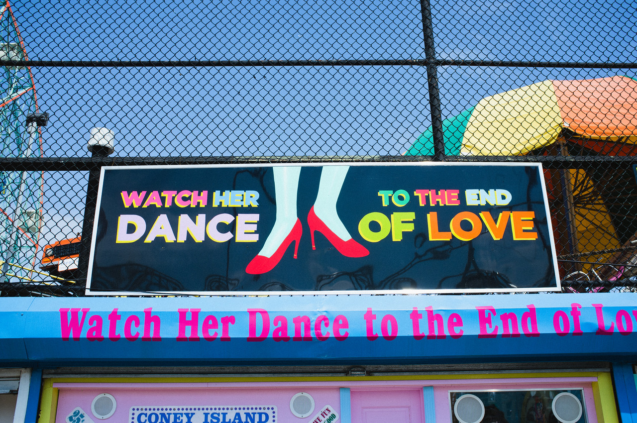 Coney Island sign reading 'WATCH HER DANCE TO THE END OF LOVE' illustrated with red high-heeled shoes.