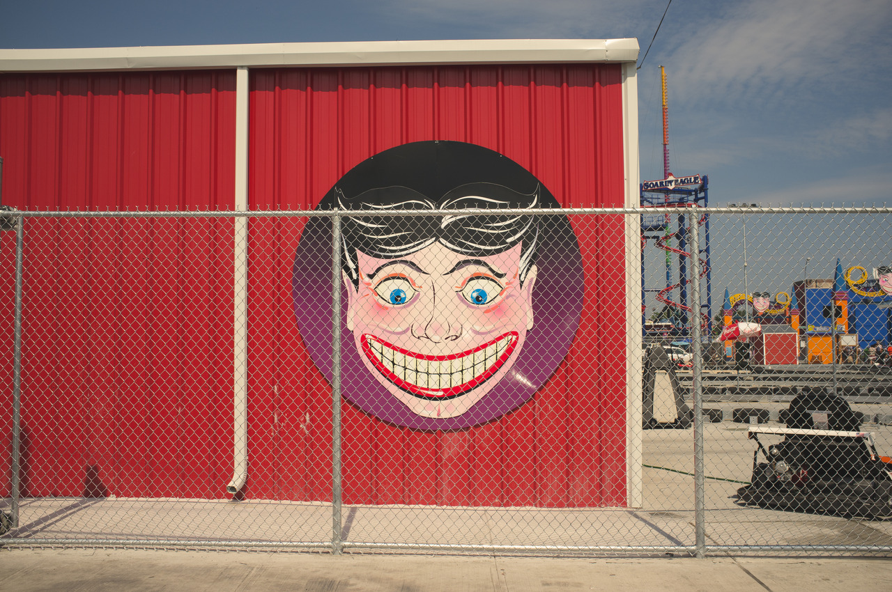 Smiling Steeplechase Face on red corrugated siding behind a chain-link fence at Coney Island.