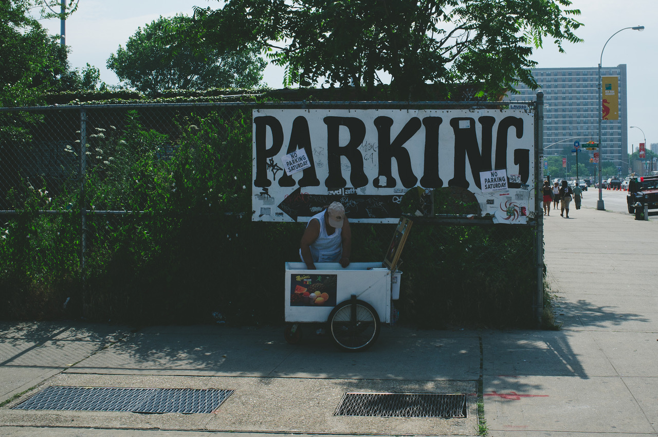 Street vendor with an Italian ice cart on a Coney Island sidewalk beside a hand-painted 'PARKING' sign.