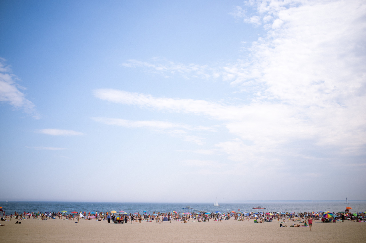 Crowded sandy beach at Coney Island filled with colorful umbrellas and sunbathers under a sunny blue sky.
