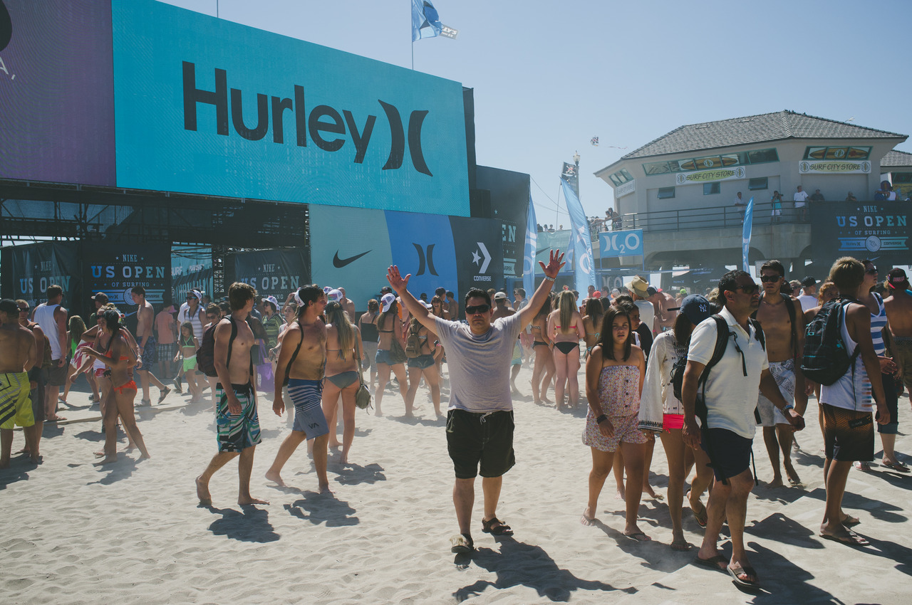 Man standing with arms outstretched on a crowded beach with large Hurley and Nike US Open of Surfing installations.