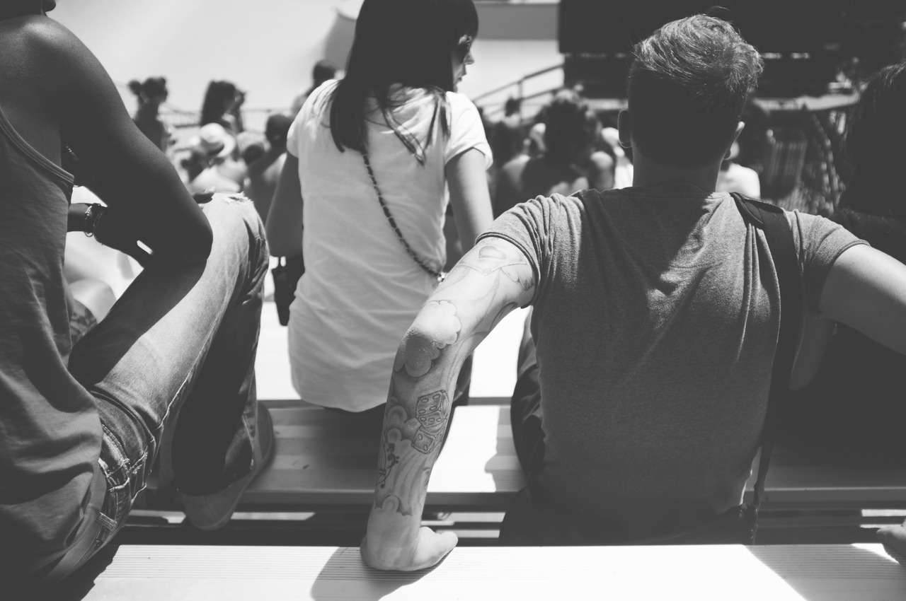 Spectators sitting on bleachers in bright sunshine, with a tattooed arm resting on the bench.