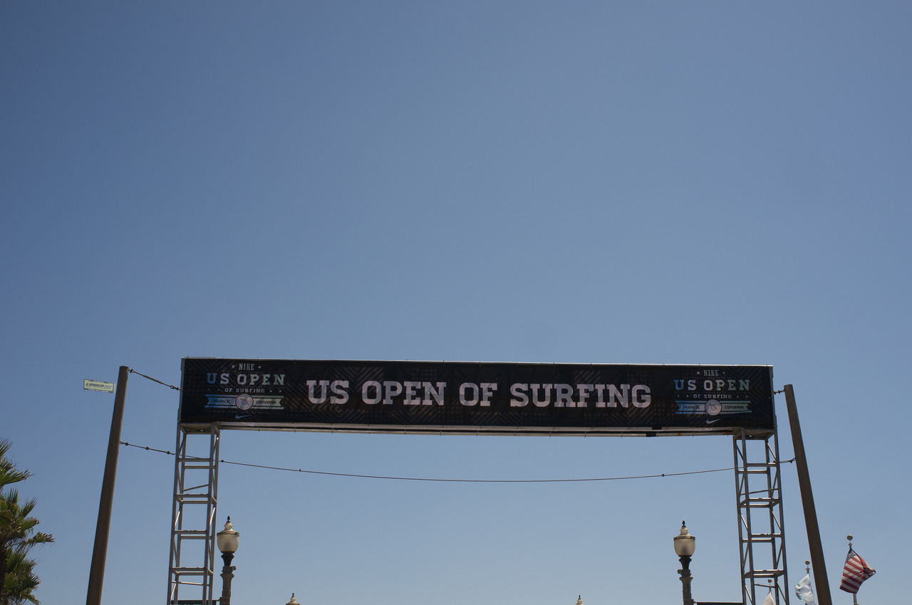 Overhead truss banner reading 'US OPEN OF SURFING' suspended between streetlamps against a clear California sky.