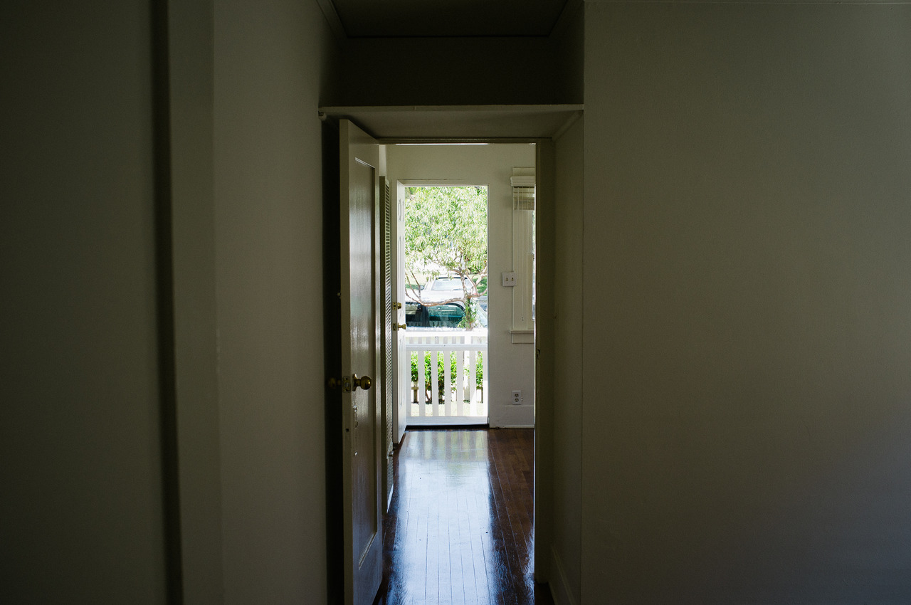 View down an interior hallway through an open doorway toward a sunlit front porch and leafy green trees.