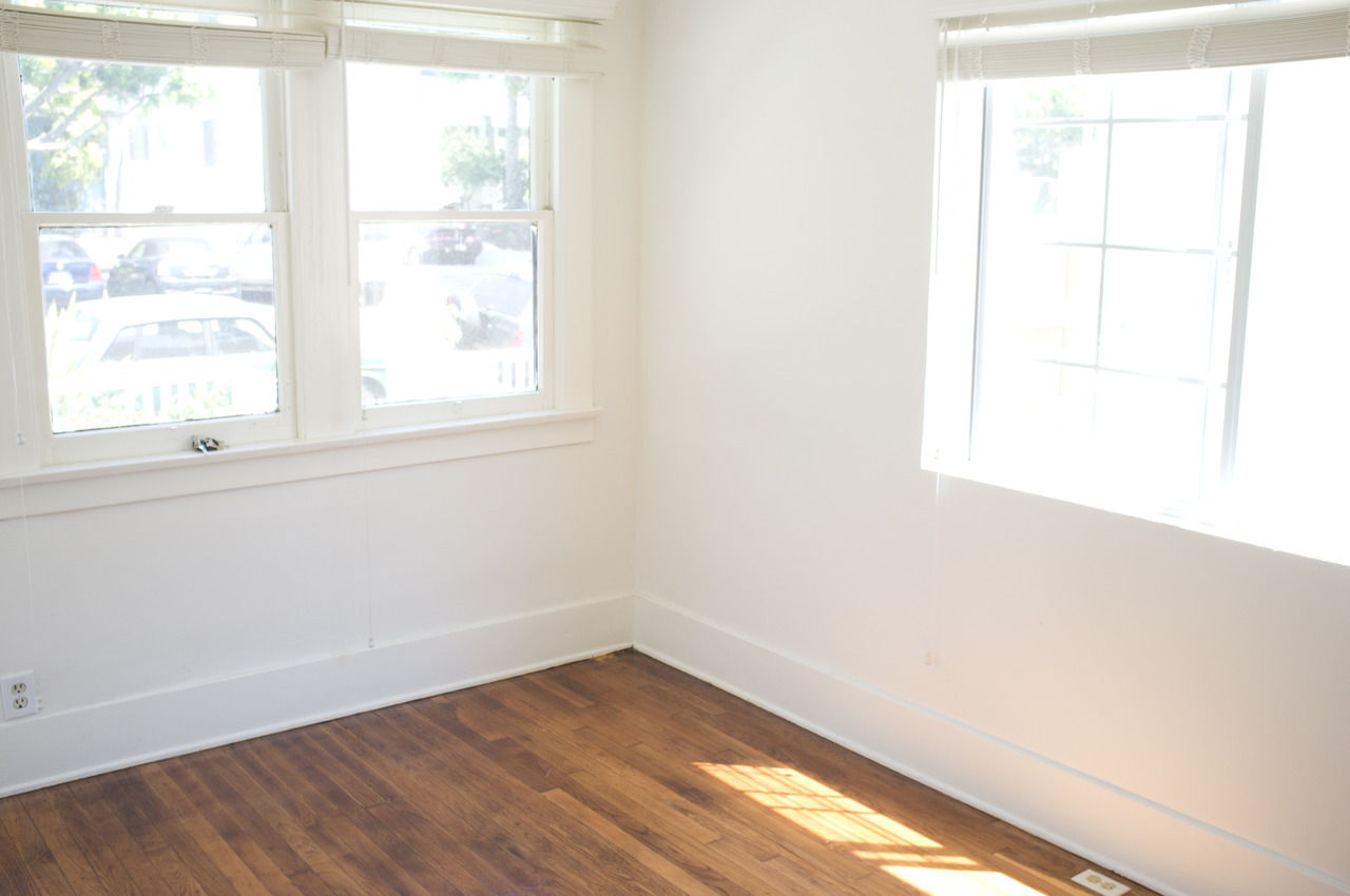 Empty apartment room corner with polished hardwood floors and bright white walls lit by natural daylight.
