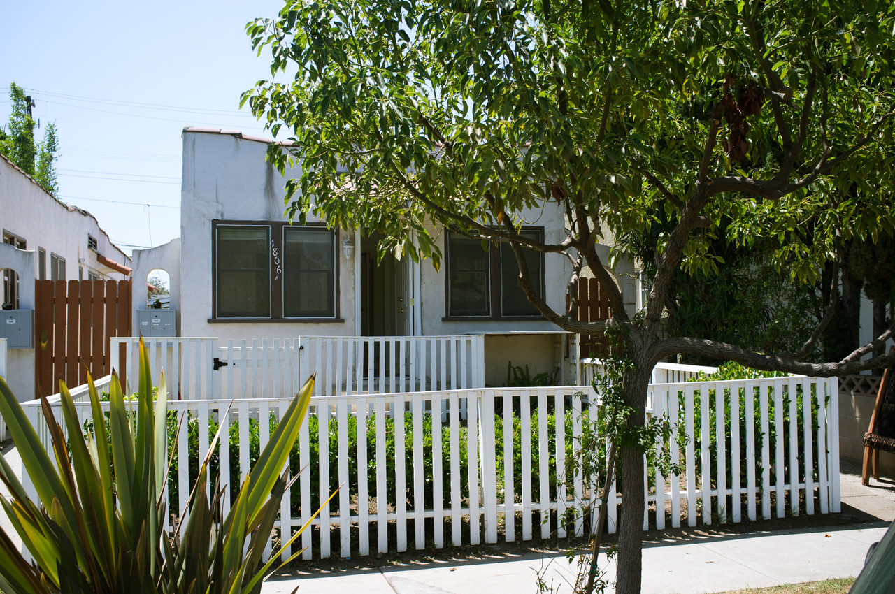 White Spanish-style bungalow behind a white picket fence and green hedges beneath leafy tree branches.