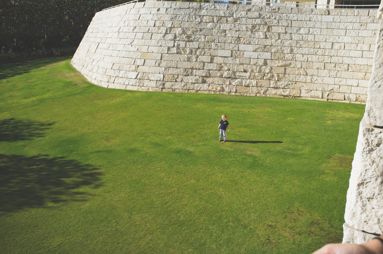 Toddler boy standing on a vast green lawn beneath a towering curved travertine stone wall at the Getty Center, captured with a Fujifilm FinePix X100.