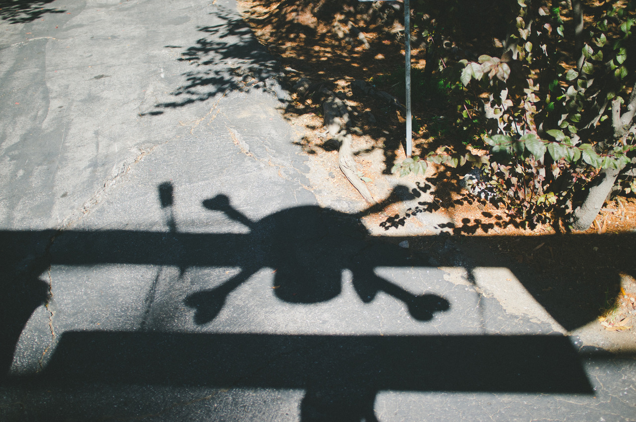 Shadow of a wooden post with a skull and crossbones silhouette cast across asphalt pavement in Oak Glen, captured with a Fujifilm FinePix X100.
