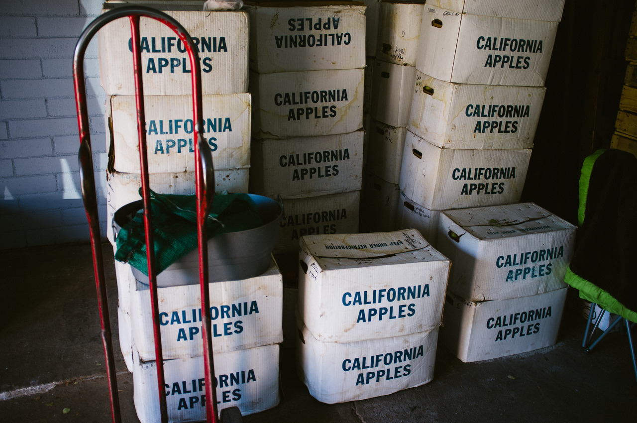 Stacks of white cardboard produce crates printed with 'CALIFORNIA APPLES' next to a red hand truck at an Oak Glen orchard, captured with a Fujifilm FinePix X100.