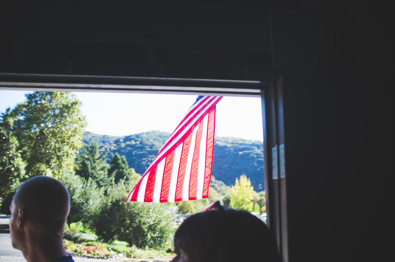 American flag fluttering outside an open doorway overlooking sunlit green hills in Oak Glen, captured with a Fujifilm FinePix X100.