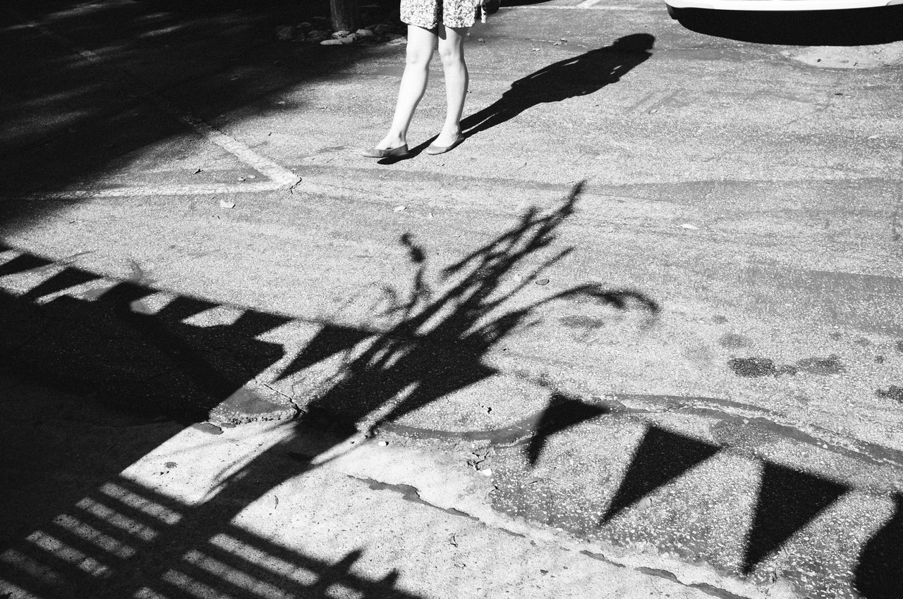 Black and white shadow of a woman walking across pavement beside shadows of decorative triangle bunting flags, captured with a Fujifilm FinePix X100.