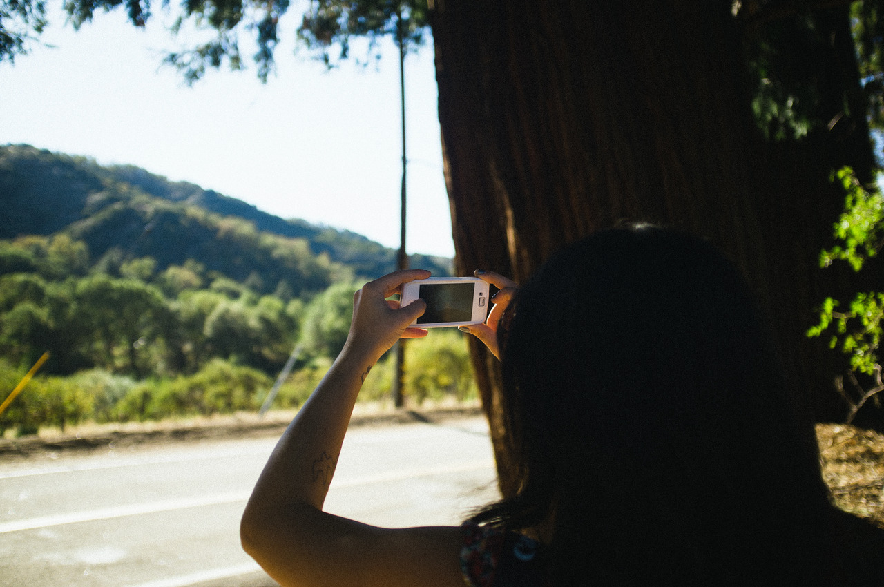 Carmen leaning against a tree trunk while photographing a mountain road with a white smartphone in Oak Glen, captured with a Fujifilm FinePix X100.