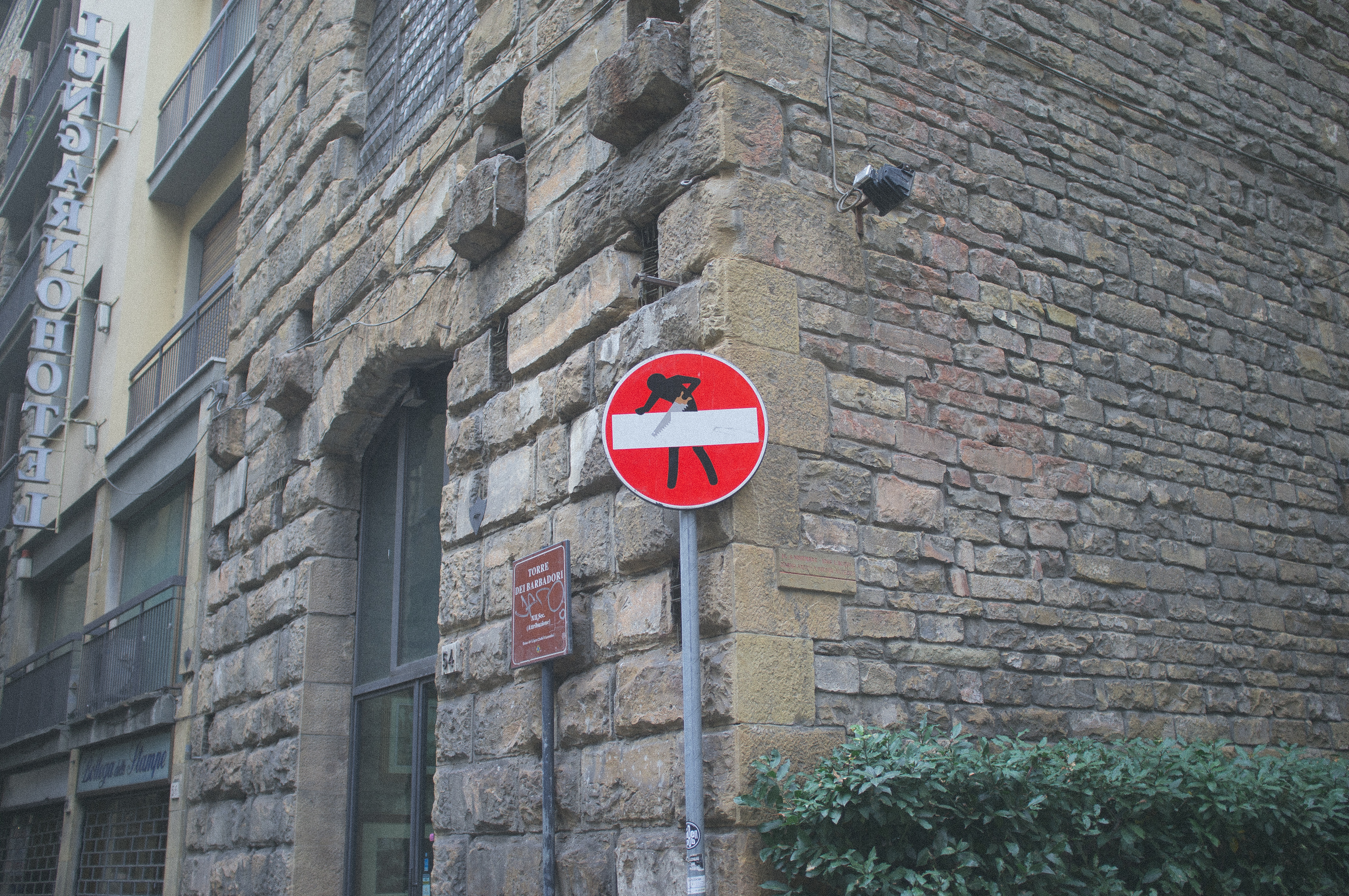 Clet Abraham street art modifying a red 'Do Not Enter' sign on a historic stone tower wall in Florence, showing a silhouette sawing through the white bar, captured with a Fujifilm FinePix X100.