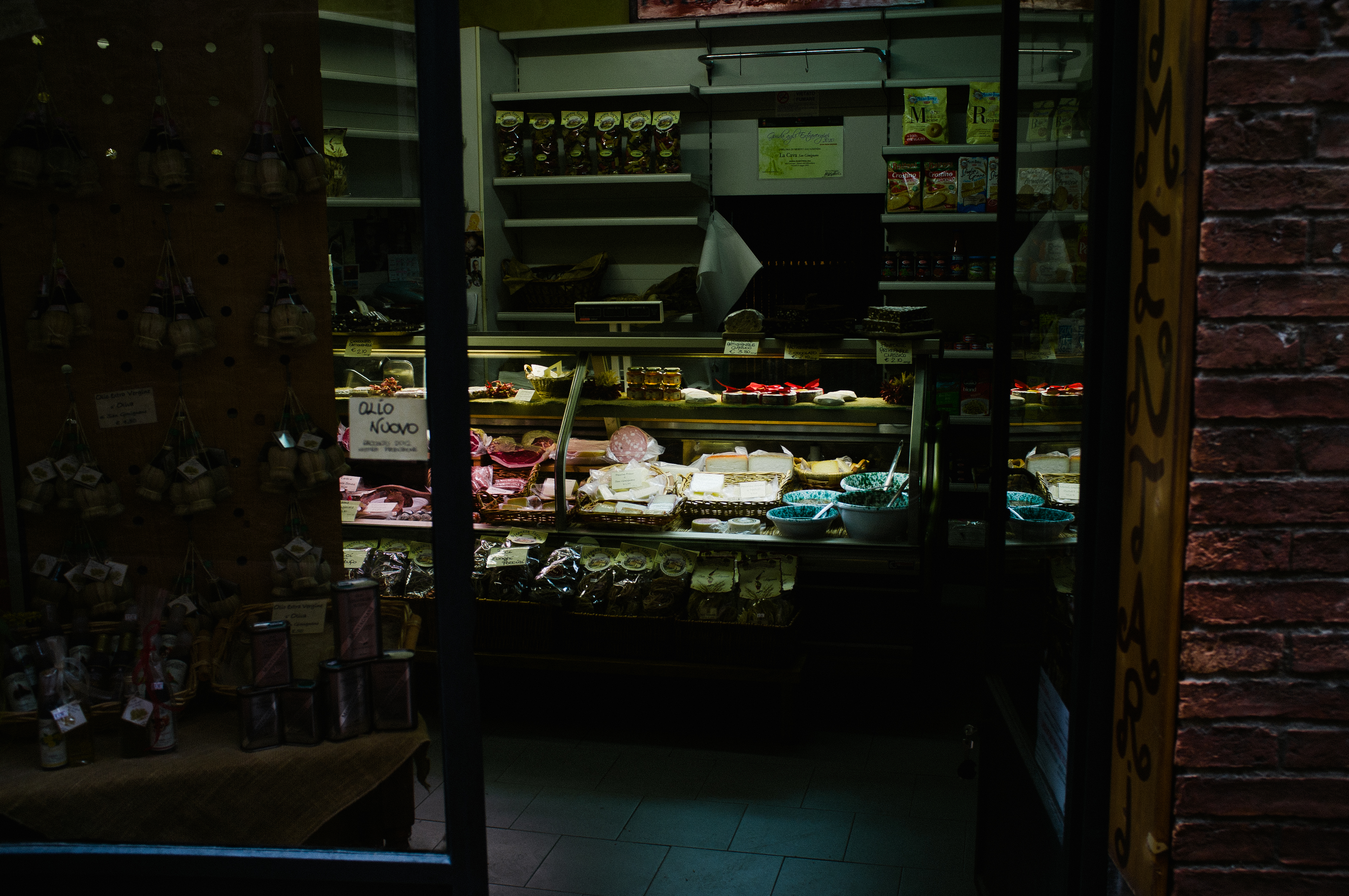 Warmly lit storefront window of an Italian deli display featuring wheels of cheese, cured meats, and an 'Olio Nuovo' sign in Florence, captured with a Fujifilm FinePix X100.