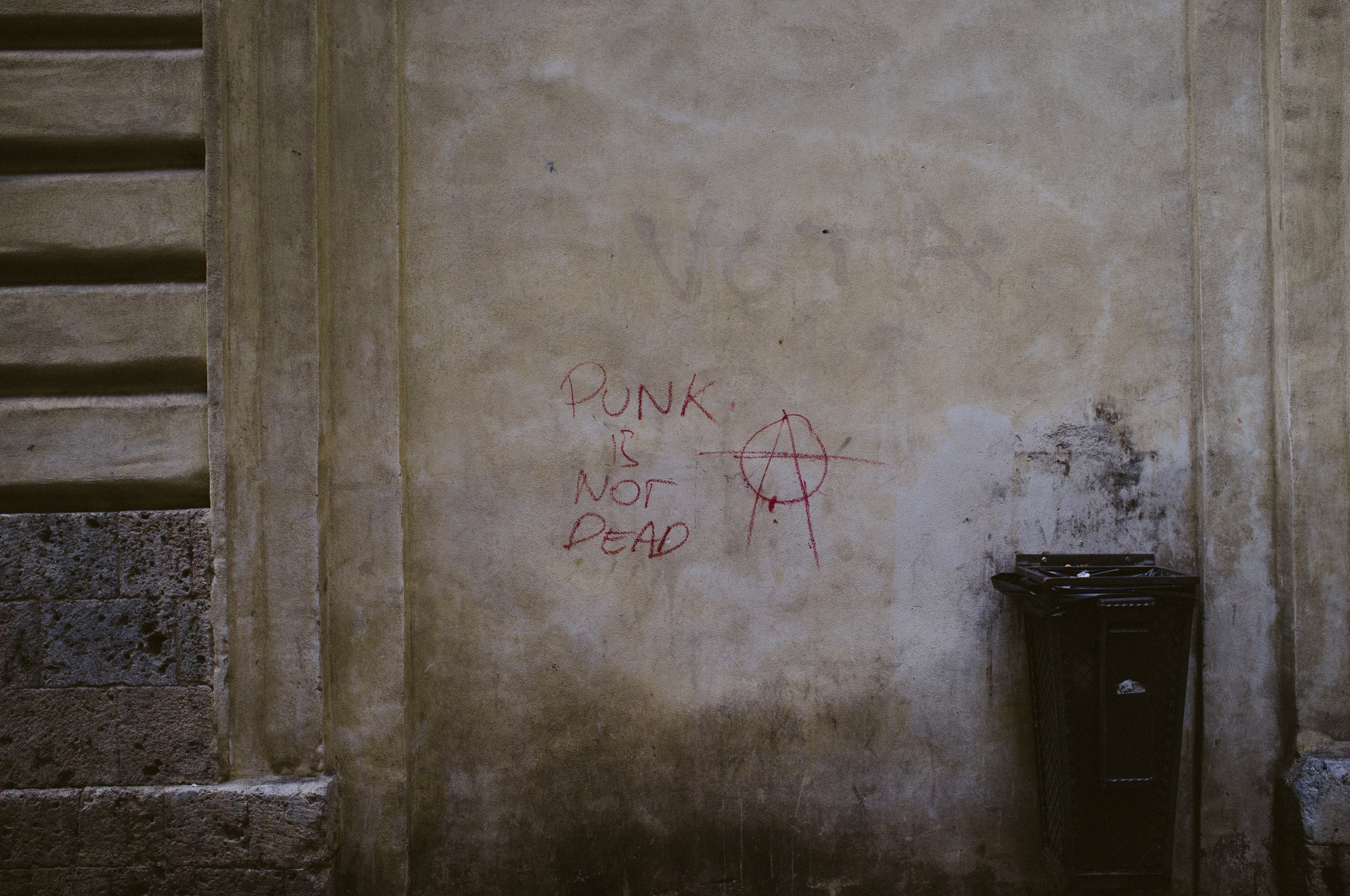 Red spray-painted graffiti reading 'PUNK IS NOT DEAD' with an anarchy symbol on a textured Italian street wall, captured with a Fujifilm FinePix X100.