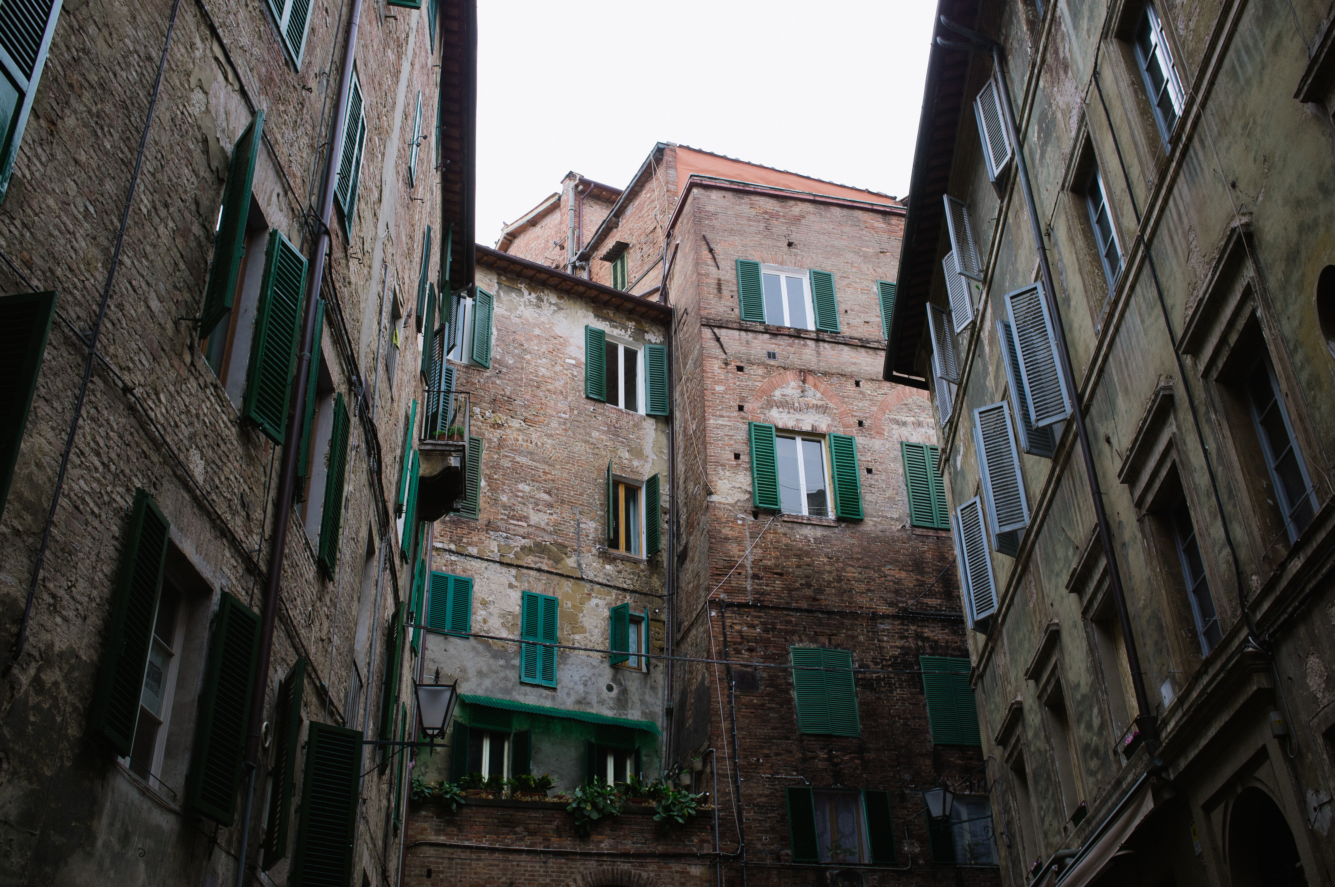 Low-angle view looking up a narrow historic alleyway between tall buildings with green window shutters in Italy.