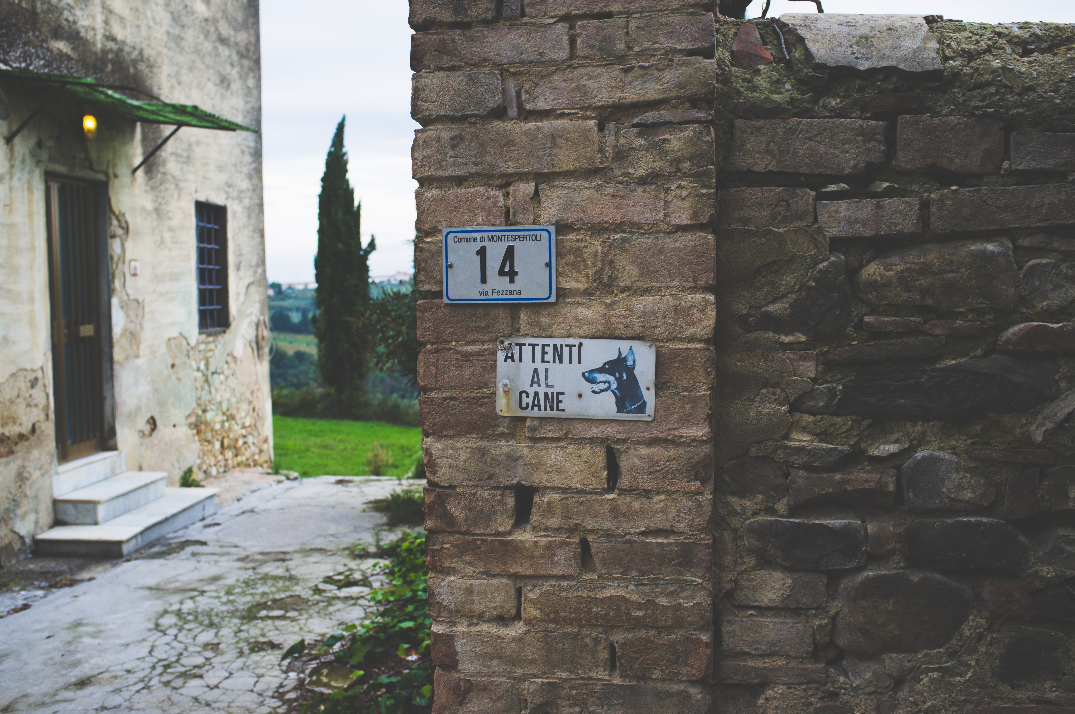 Stone wall in Tuscany featuring a '14 via Fezzana' plate and an 'Attenti al Cane' dog warning sign.