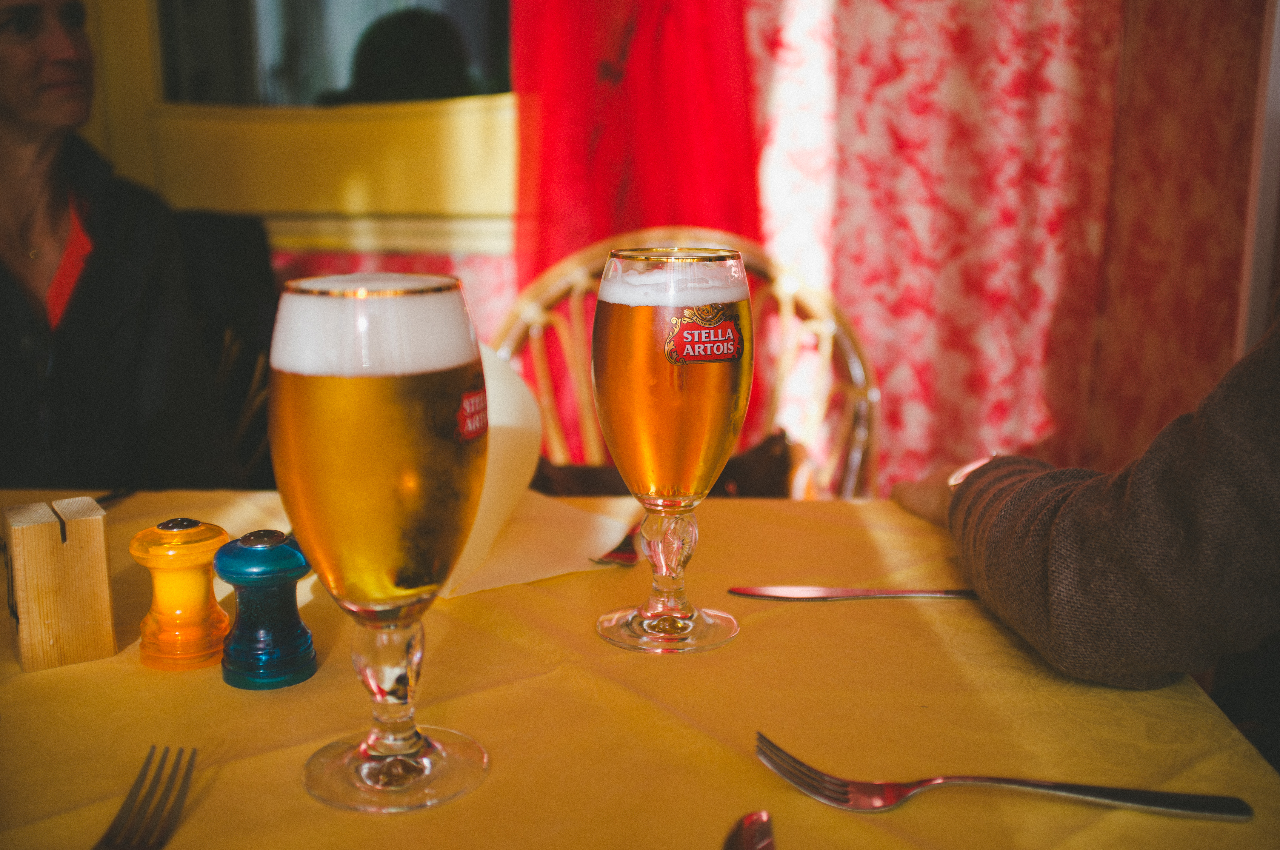 Two stemmed chalice glasses of Stella Artois beer on a restaurant table with yellow cloth and salt shakers.