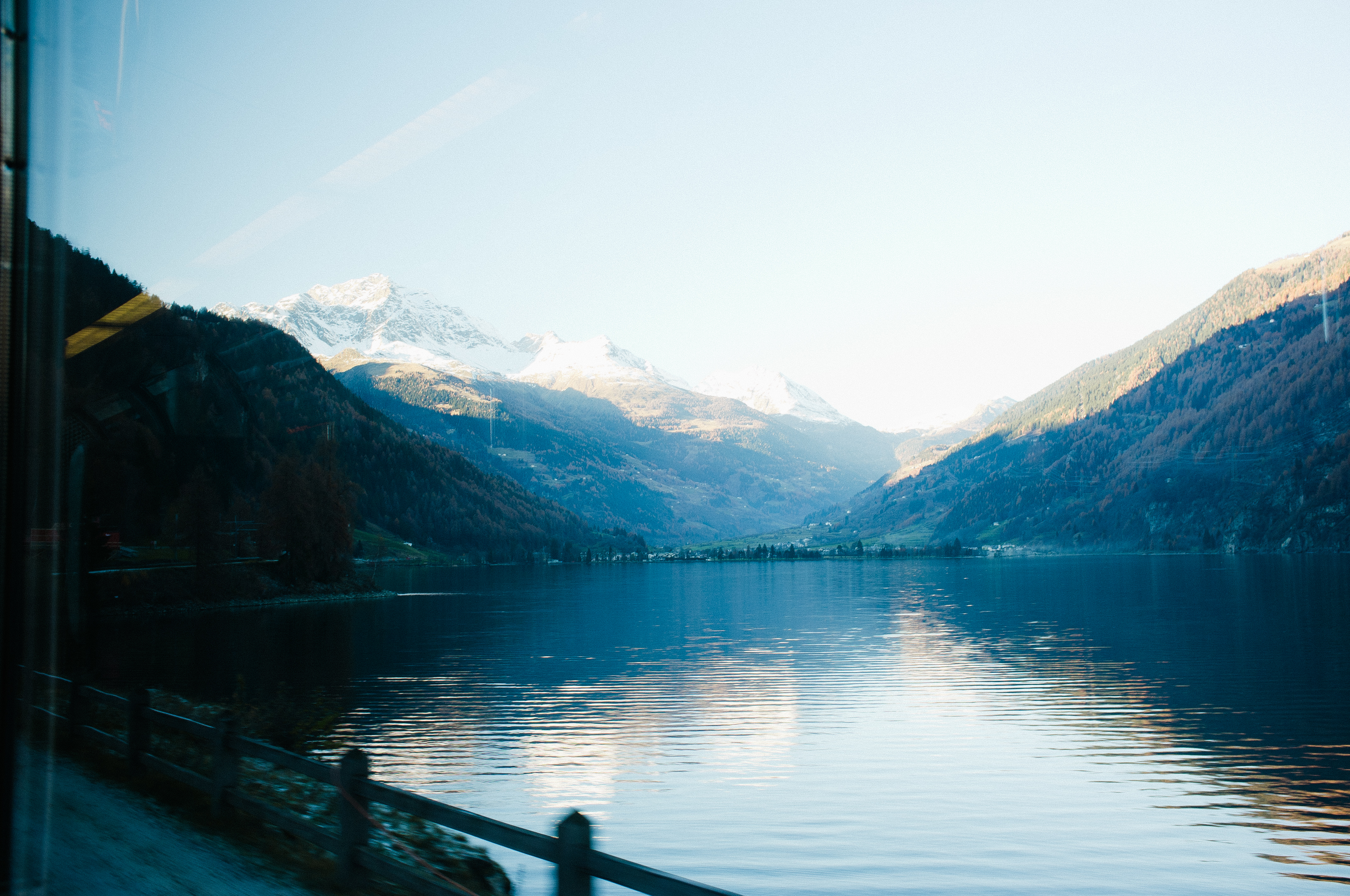 Scenic Alpine view from train window across Lake Poschiavo reflecting snow-dusted mountains and autumn foliage.