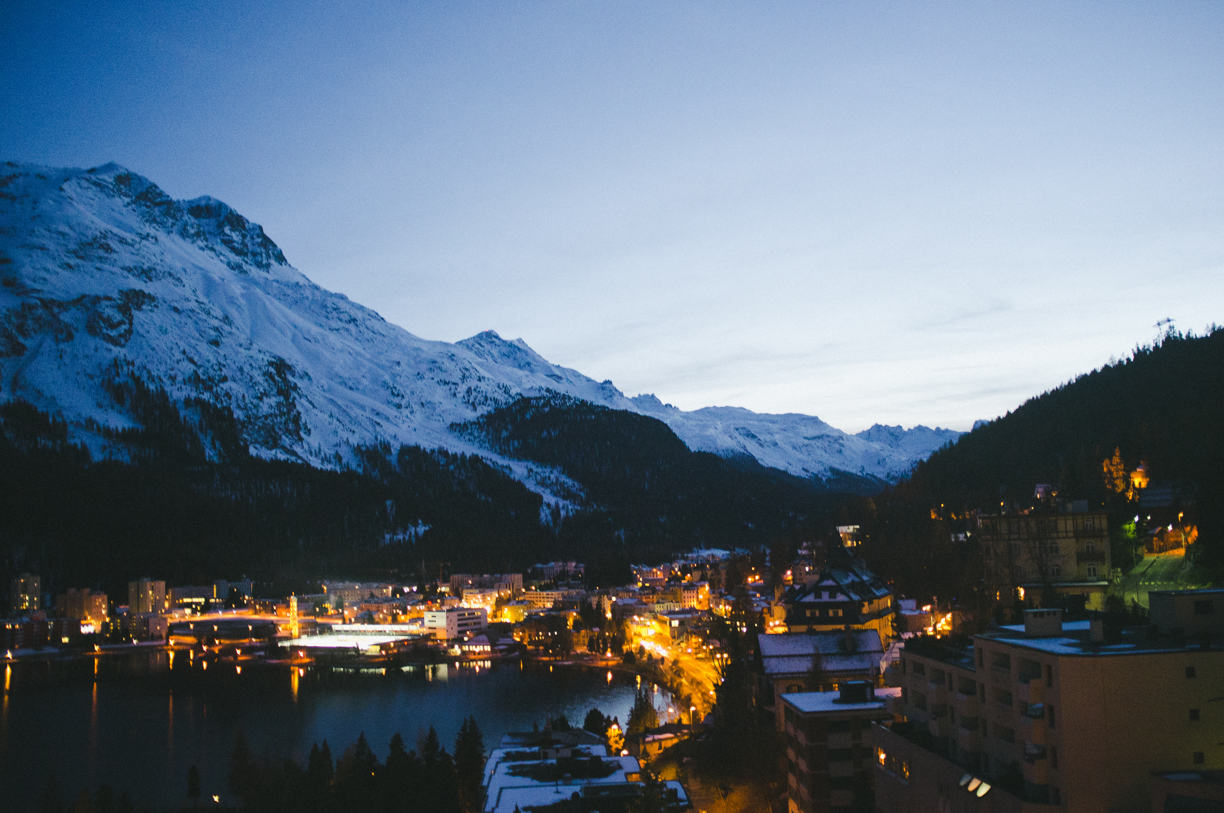 St. Moritz village illuminated along an Alpine lake shoreline beneath snowy mountain peaks at dusk.