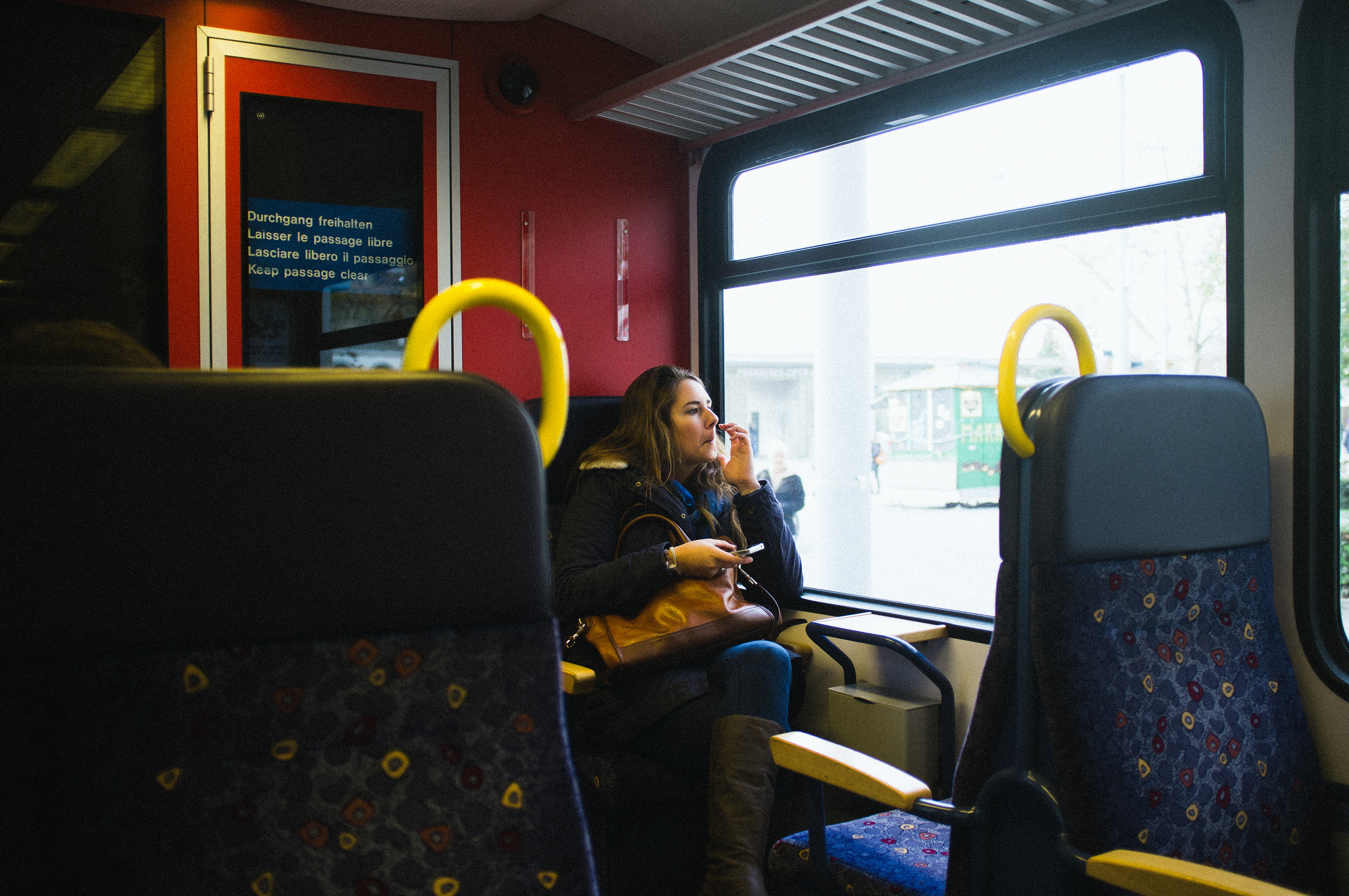 Maggie seated beside a Swiss train window holding a mobile phone to her ear with brown leather bag on lap.