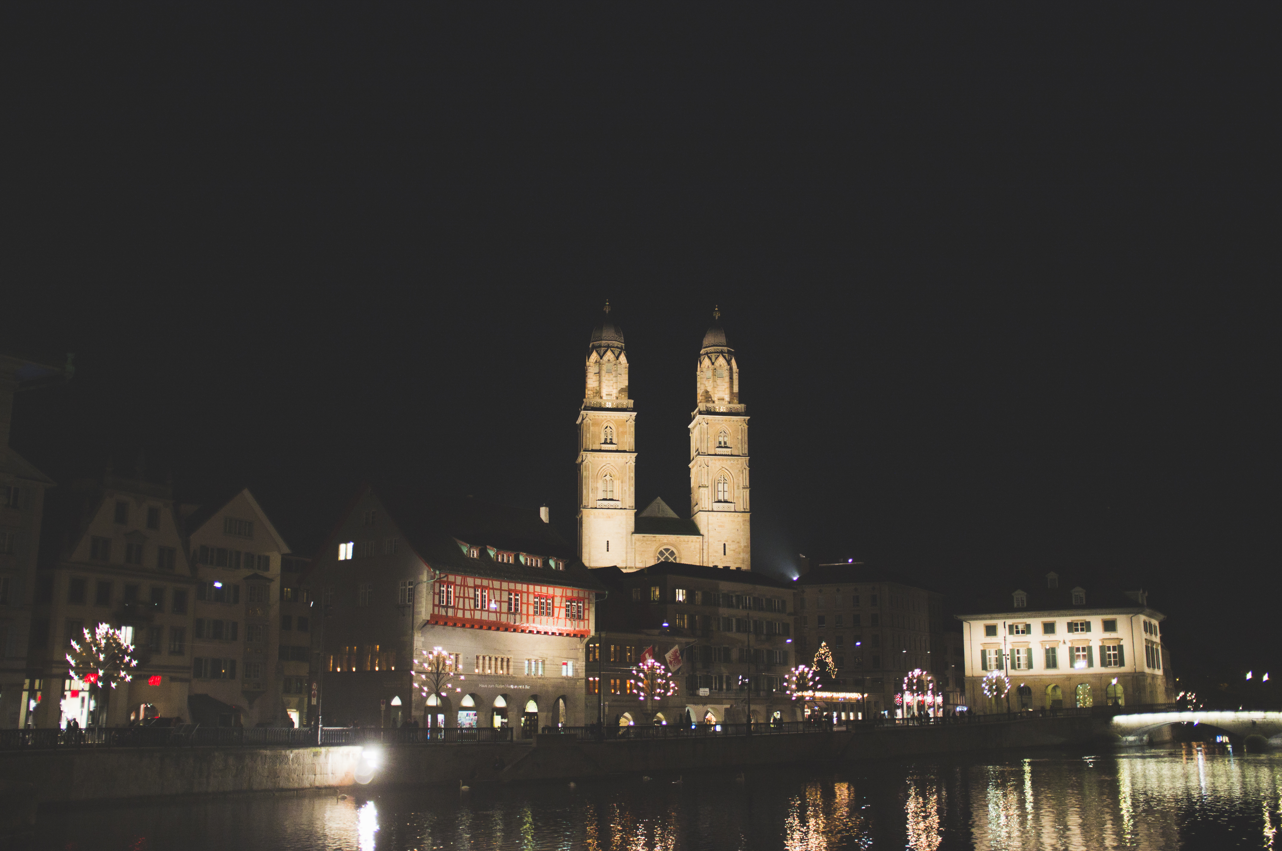 Night view of illuminated Grossmünster cathedral towers reflecting on Zurich's Limmat river.