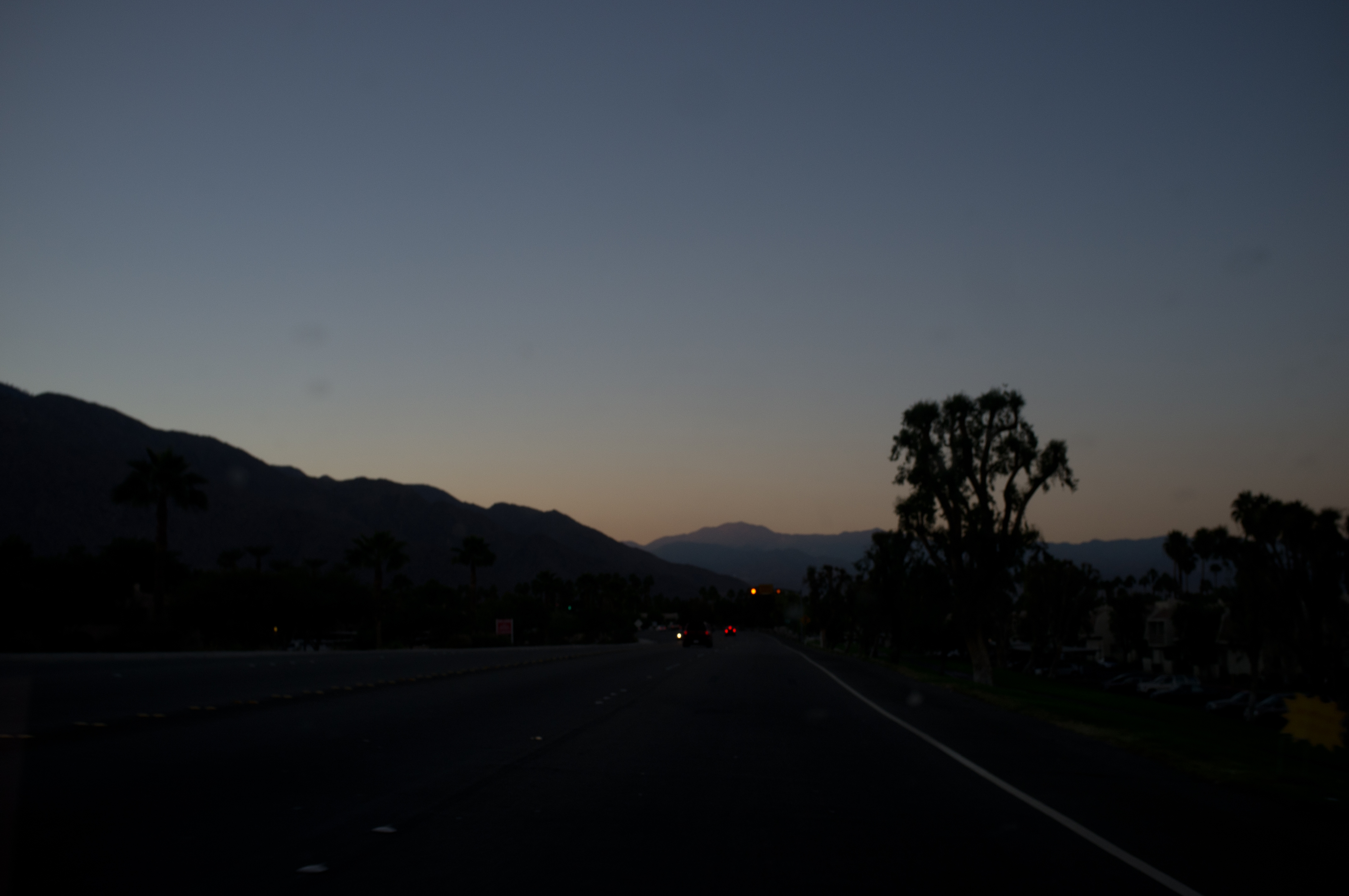 Dusk road view from a car driving toward the silhouette of Palm Springs desert mountains under deep blue twilight.