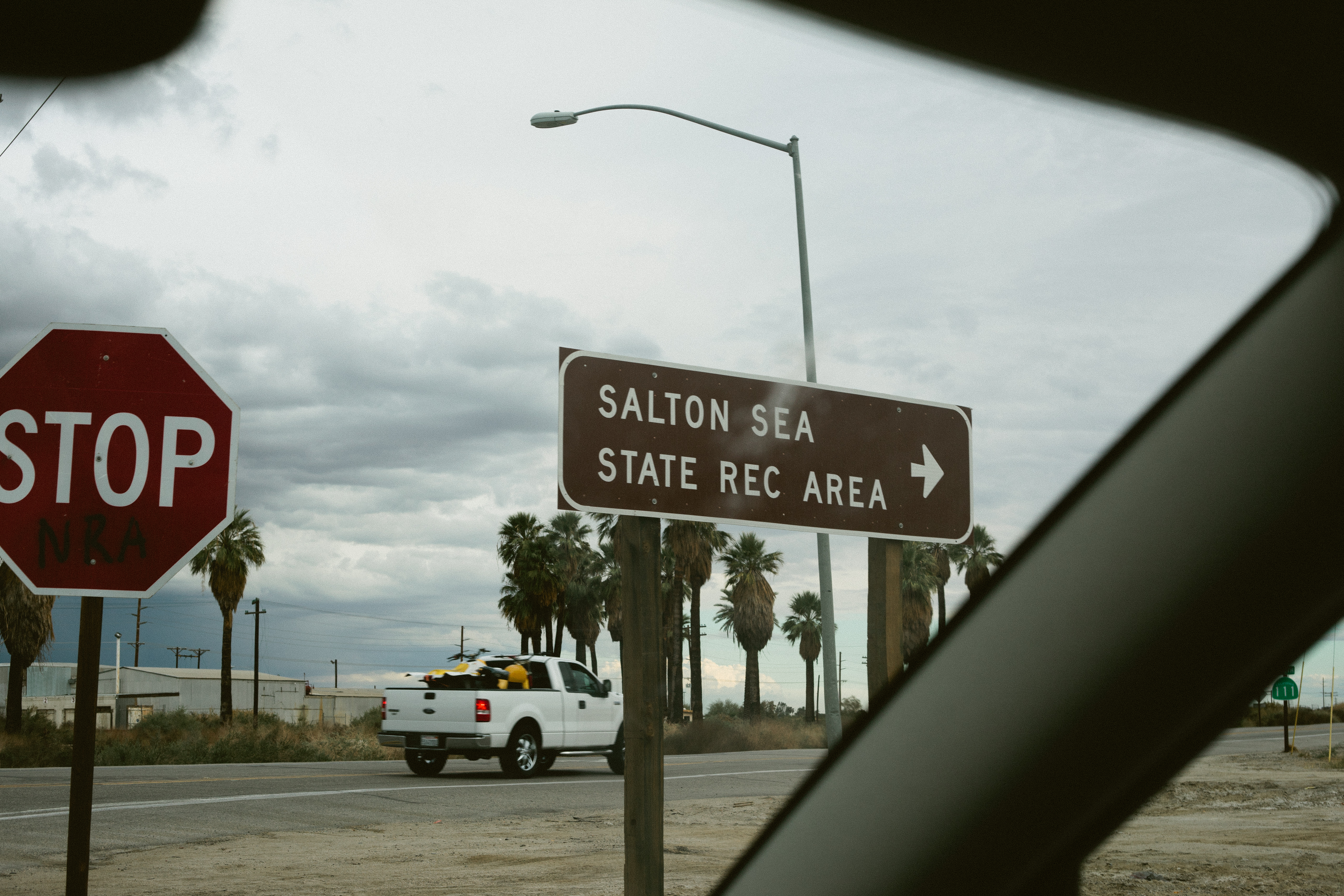Highway road trip view from a car passing a brown sign for 'SALTON SEA STATE REC AREA' beside a white pickup truck.