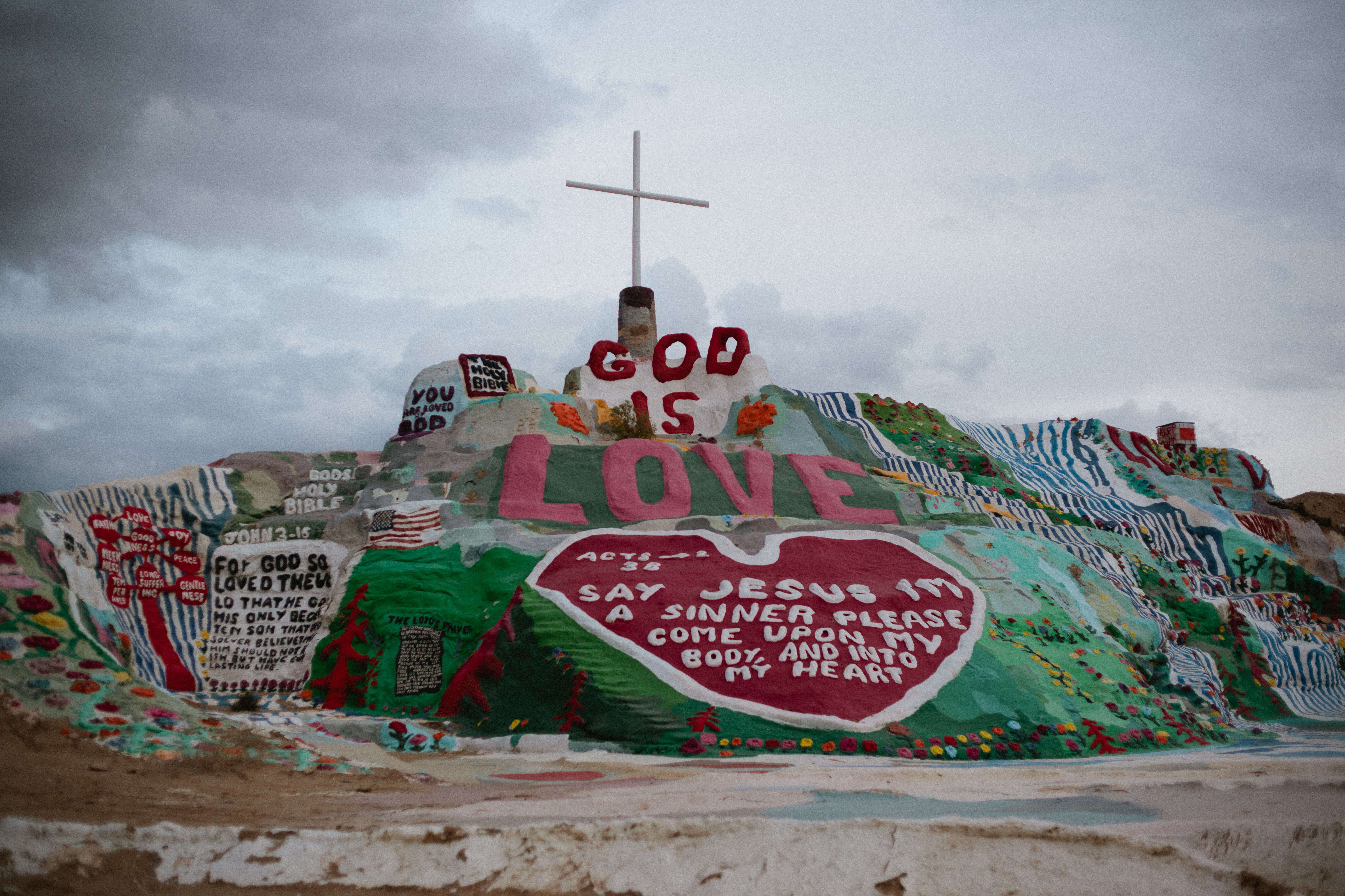 Salvation Mountain painted with a prominent white cross and 'GOD IS LOVE' under cloudy skies.