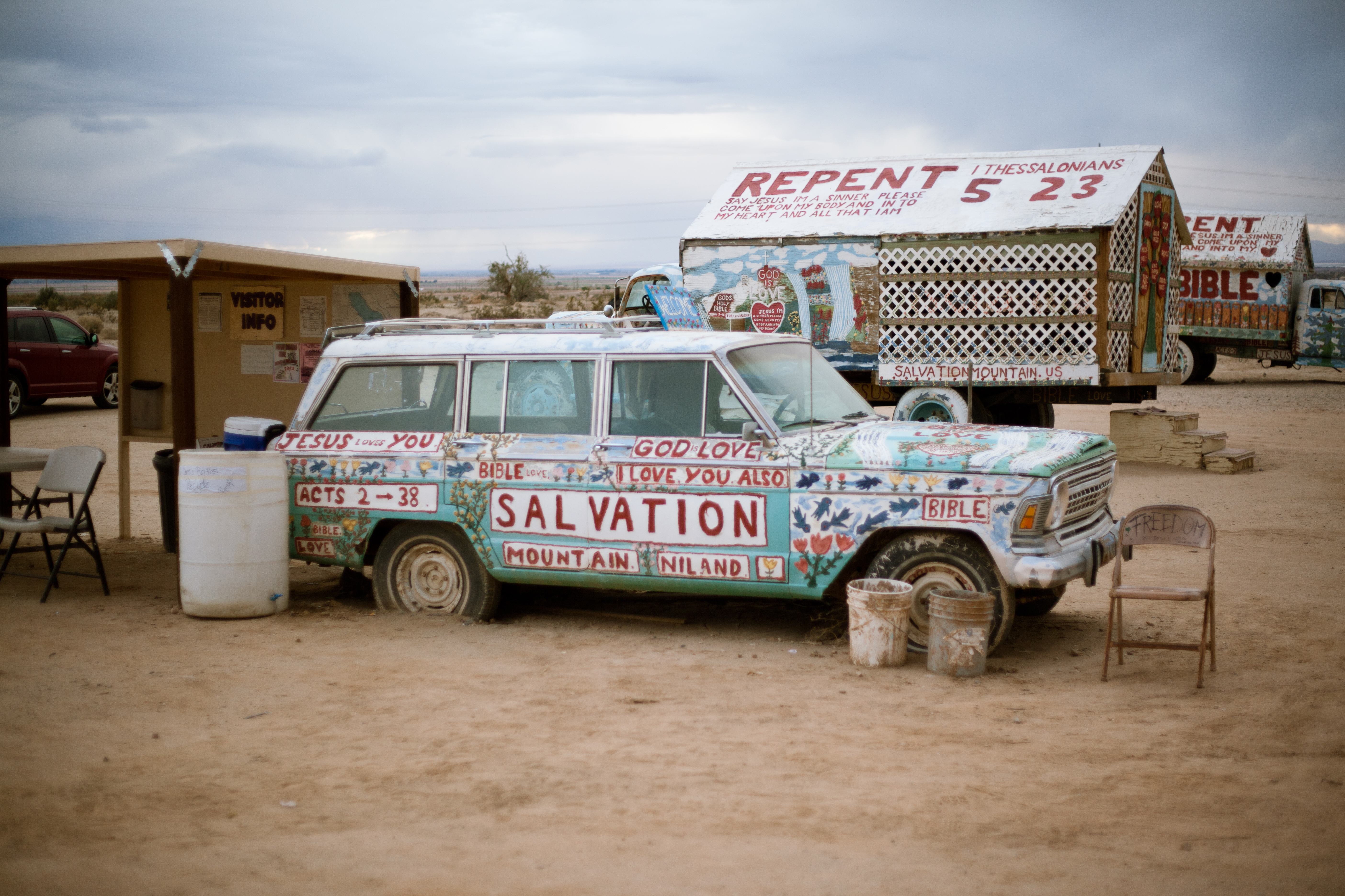 Station wagon painted with folk art reading 'SALVATION MOUNTAIN NILAND' parked in desert sand.