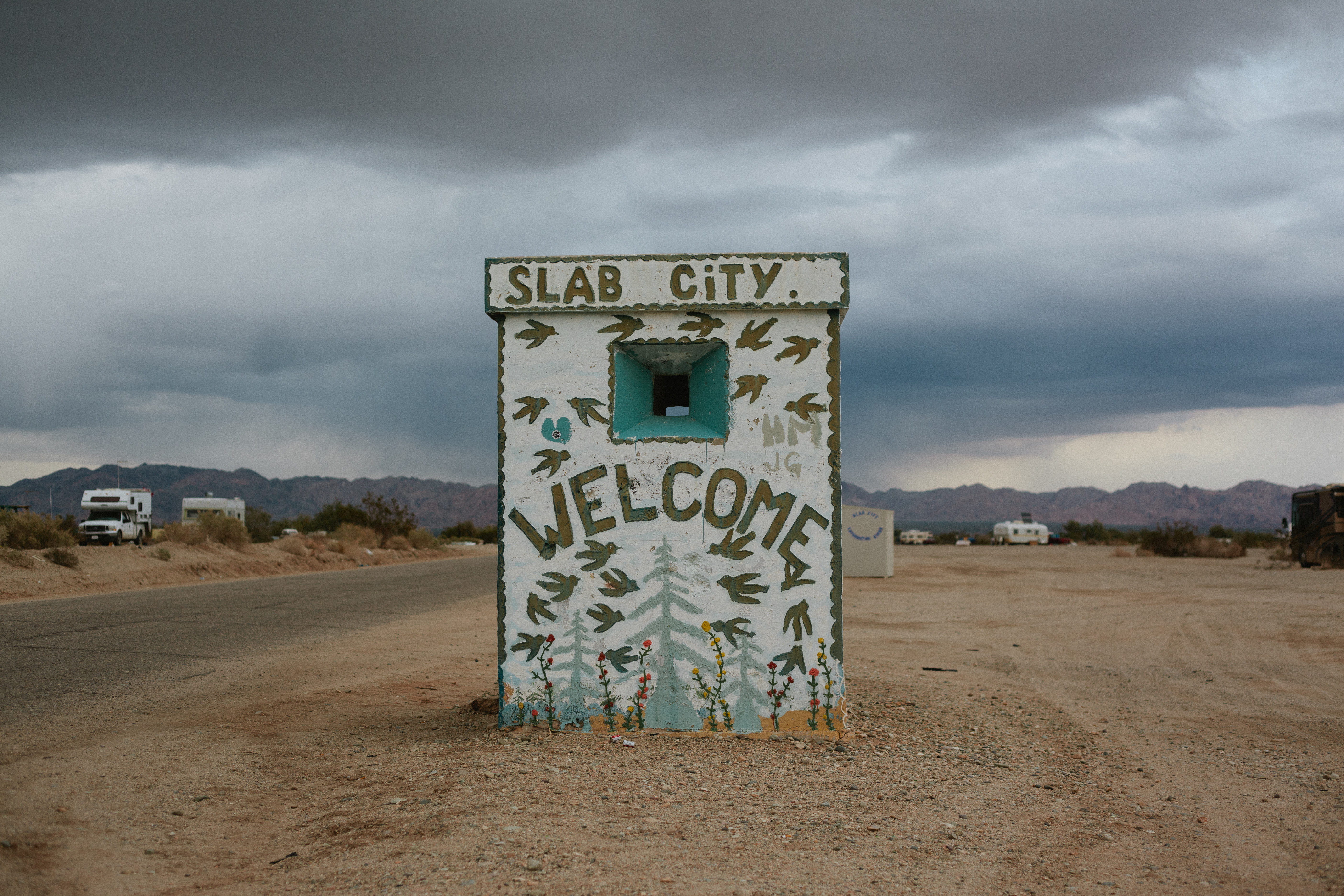 Weathered concrete guard shack painted with 'SLAB CITY' and 'WELCOME' graffiti on the dirt road entrance.